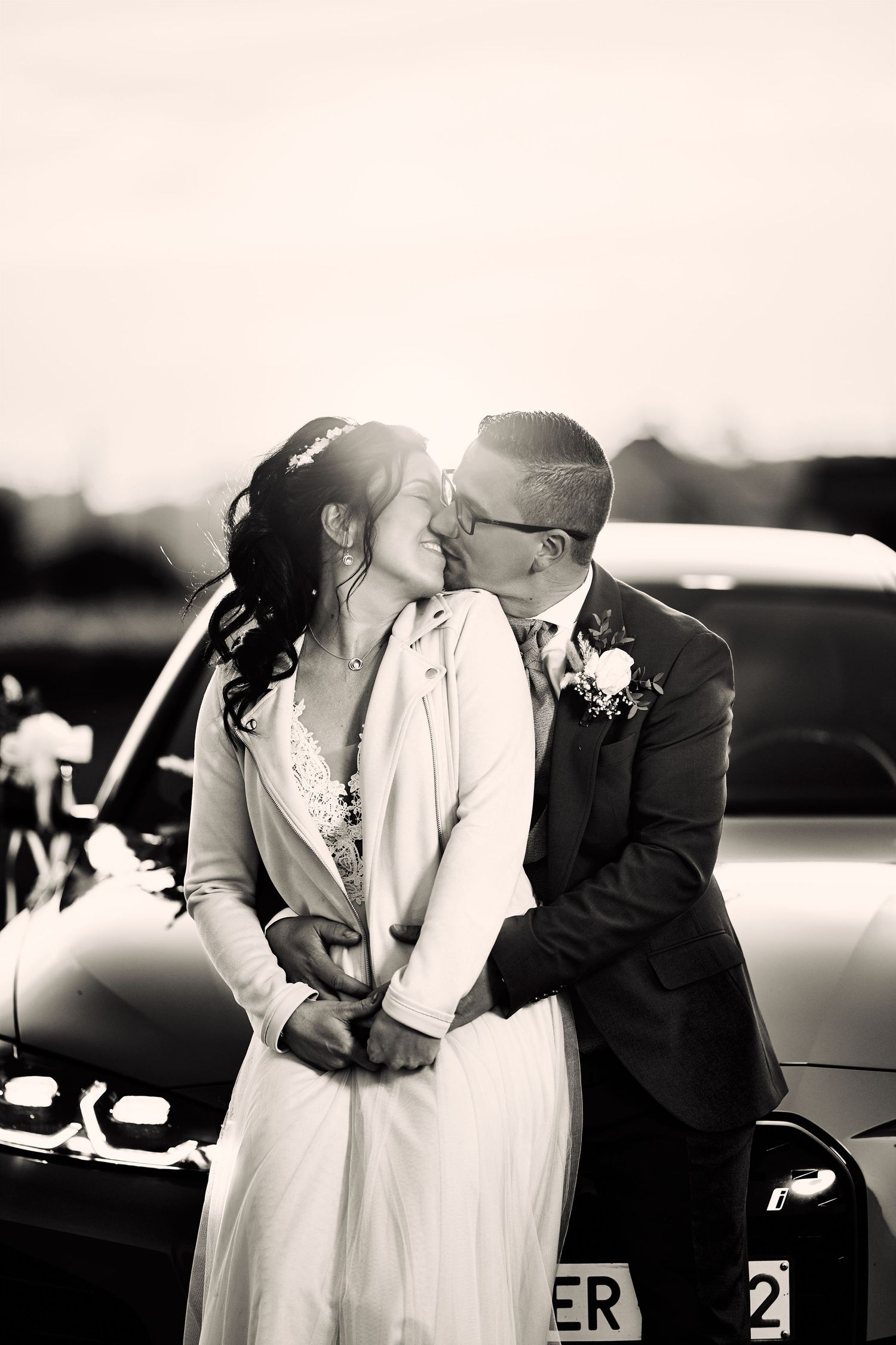 Black and white photo of a bride and groom sharing a kiss beside their decorated wedding car.