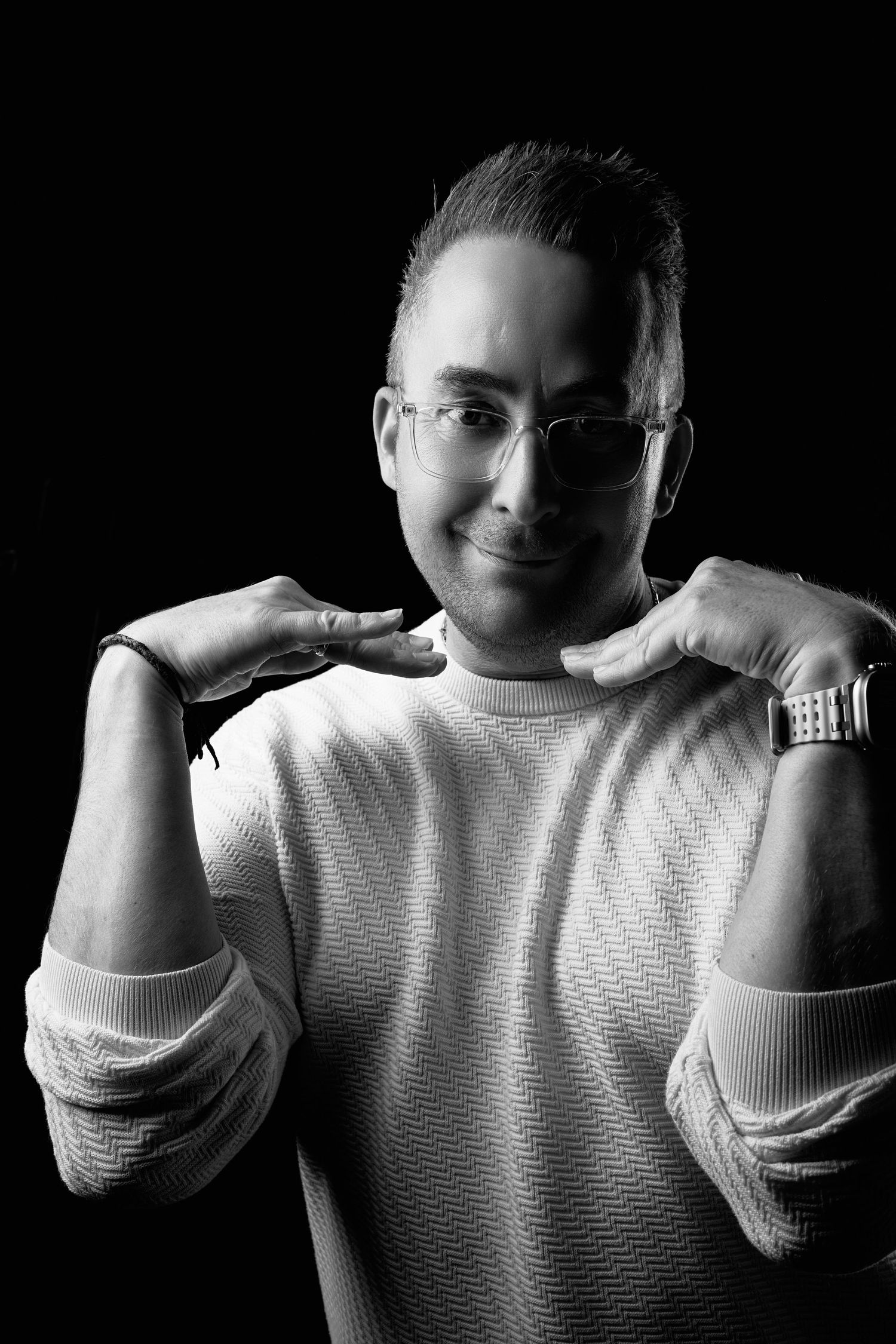 Black and white portrait of a smiling man in glasses and sweater, posing confidently with hands near shoulders.