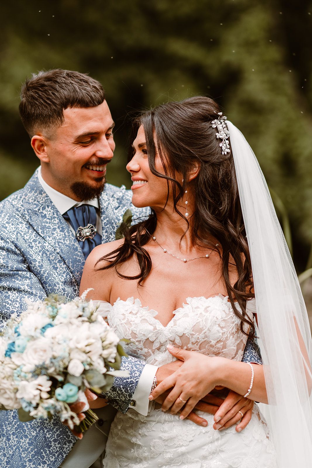 Wedding couple in silver patterned suit and white dress share intimate moment with bouquet.