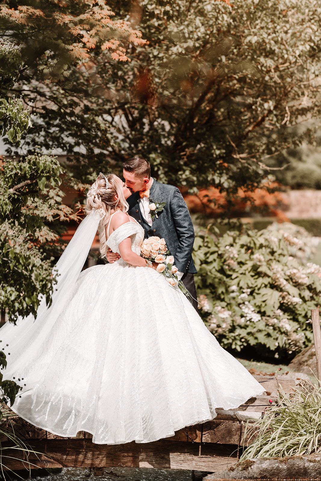 Bride in flowing white gown poses with groom on rustic wooden bench surrounded by greenery.