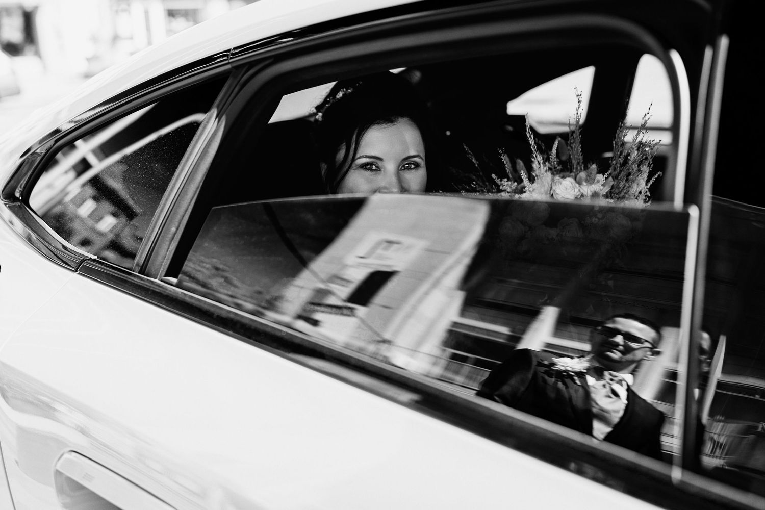 Black and white photo of a bride sitting in a car, with a photographer's reflection visible in the window.