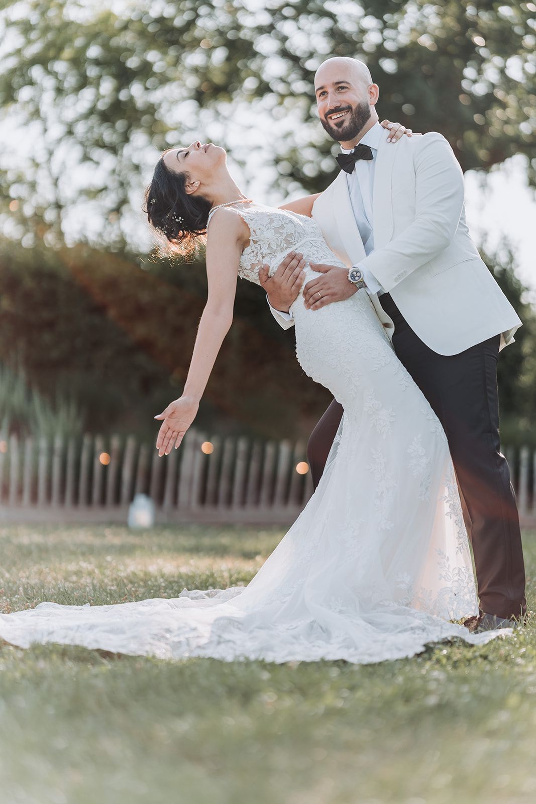 A couple in wedding attire dances and dips romantically on a grassy lawn during golden hour.