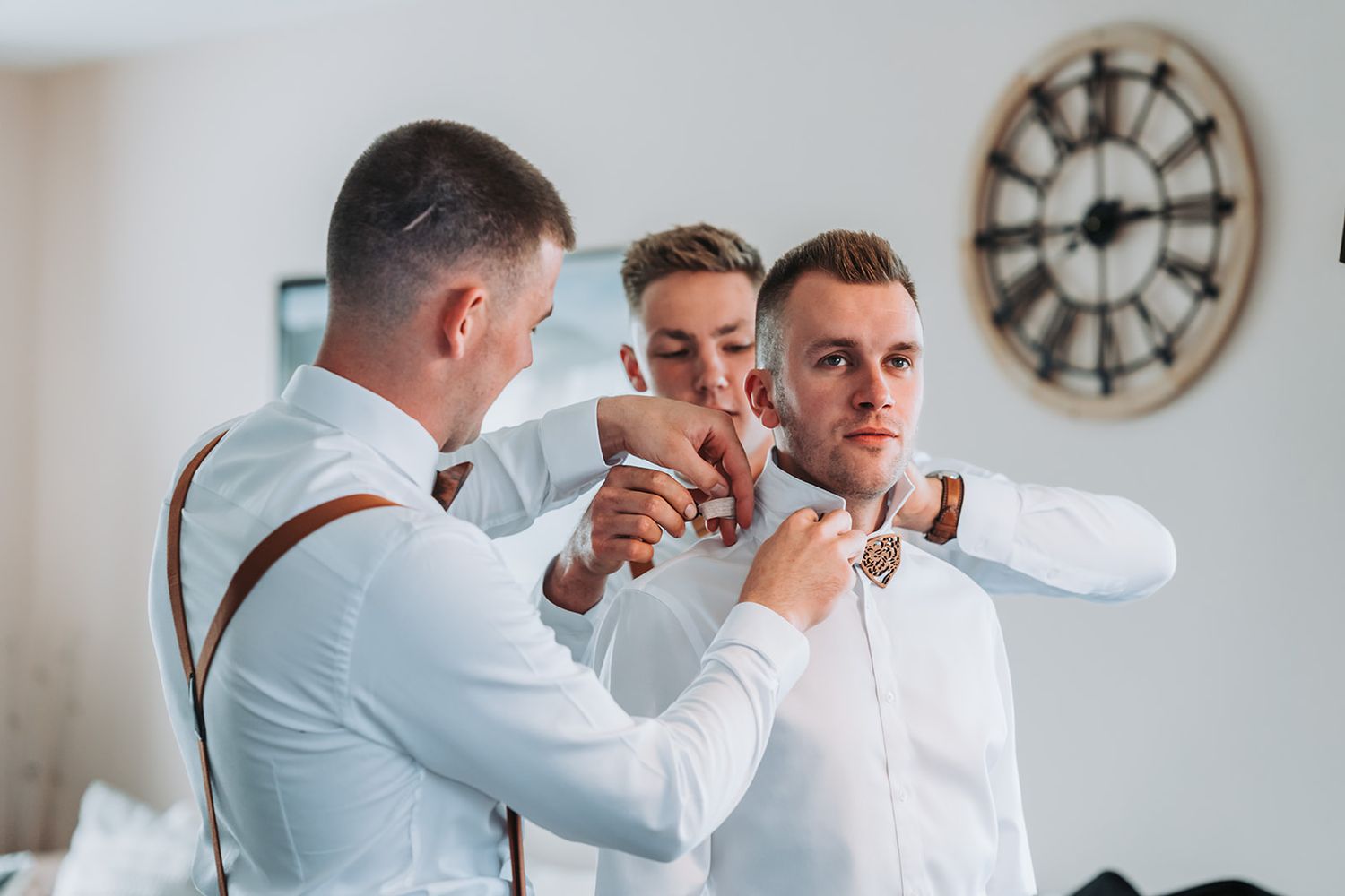 Groomsmen help with wedding preparations in room with vintage clock on wall.