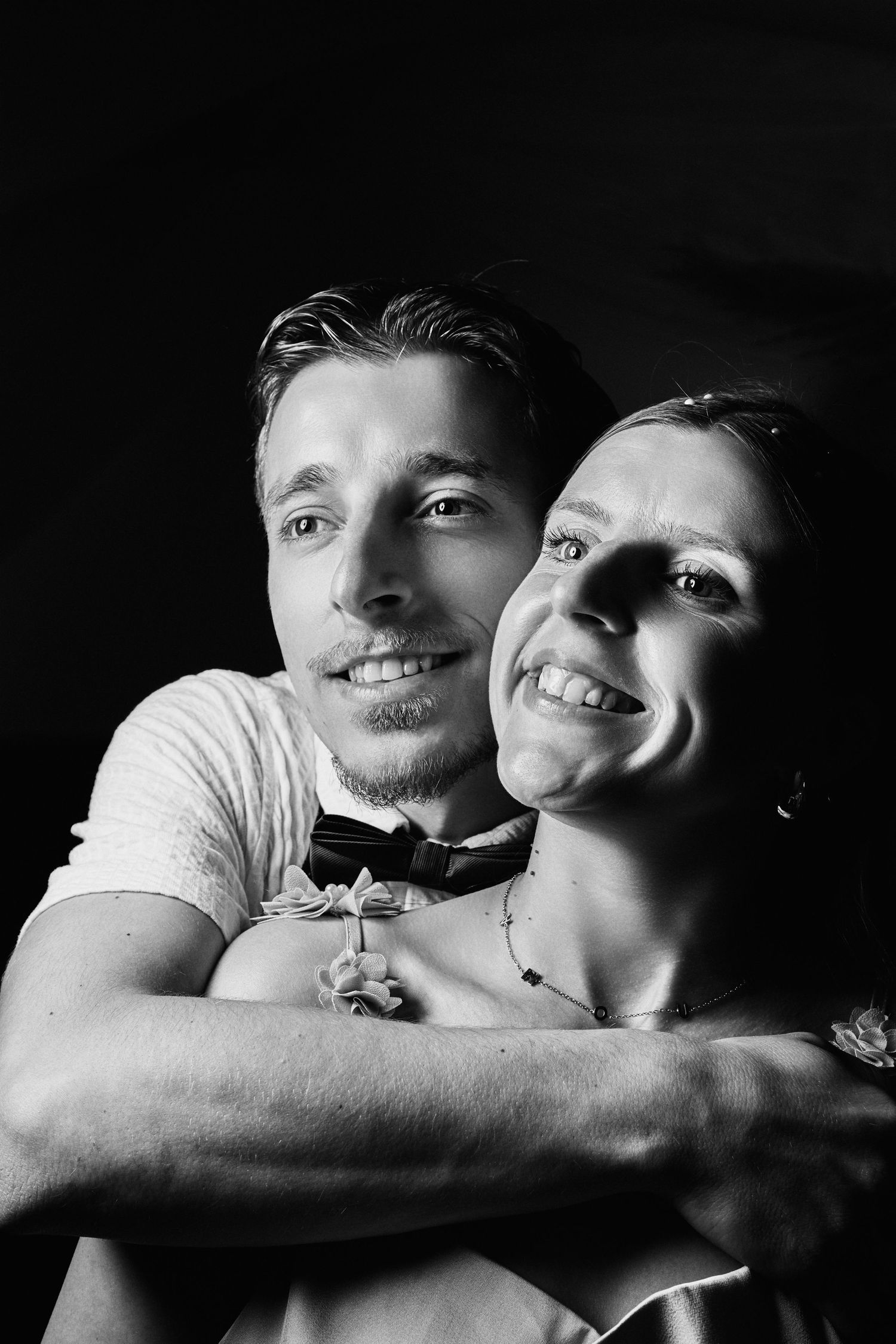 Black and white portrait of a smiling couple embracing, man in bow tie hugging woman from behind.