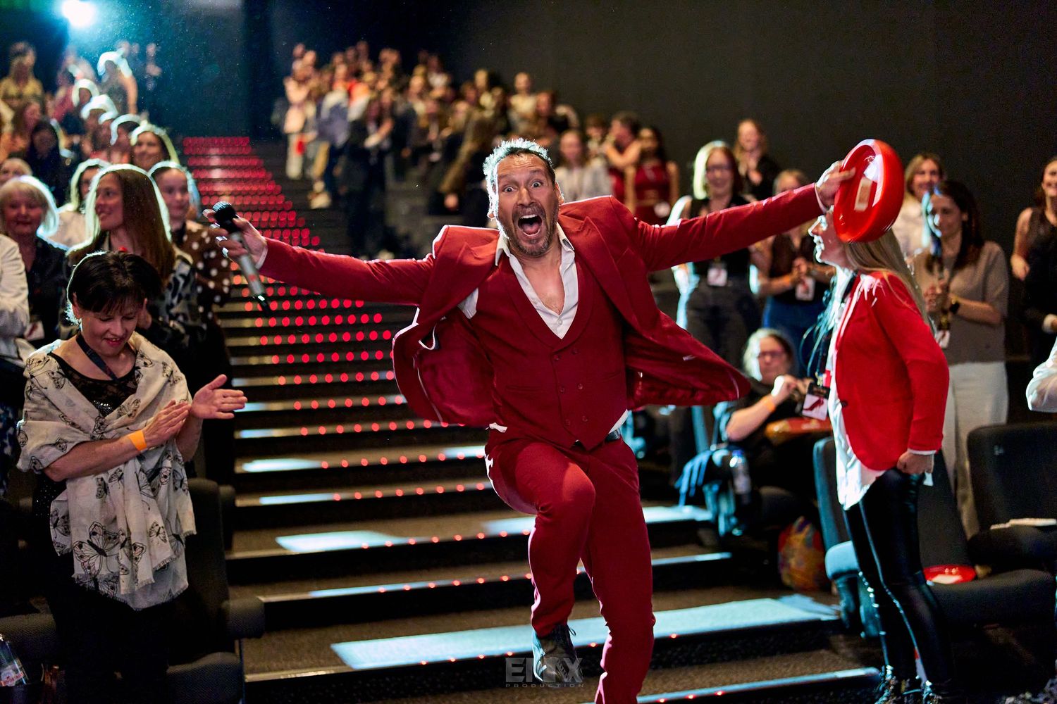 Man in red suit dancing energetically in theater aisle with audience applauding in background.
