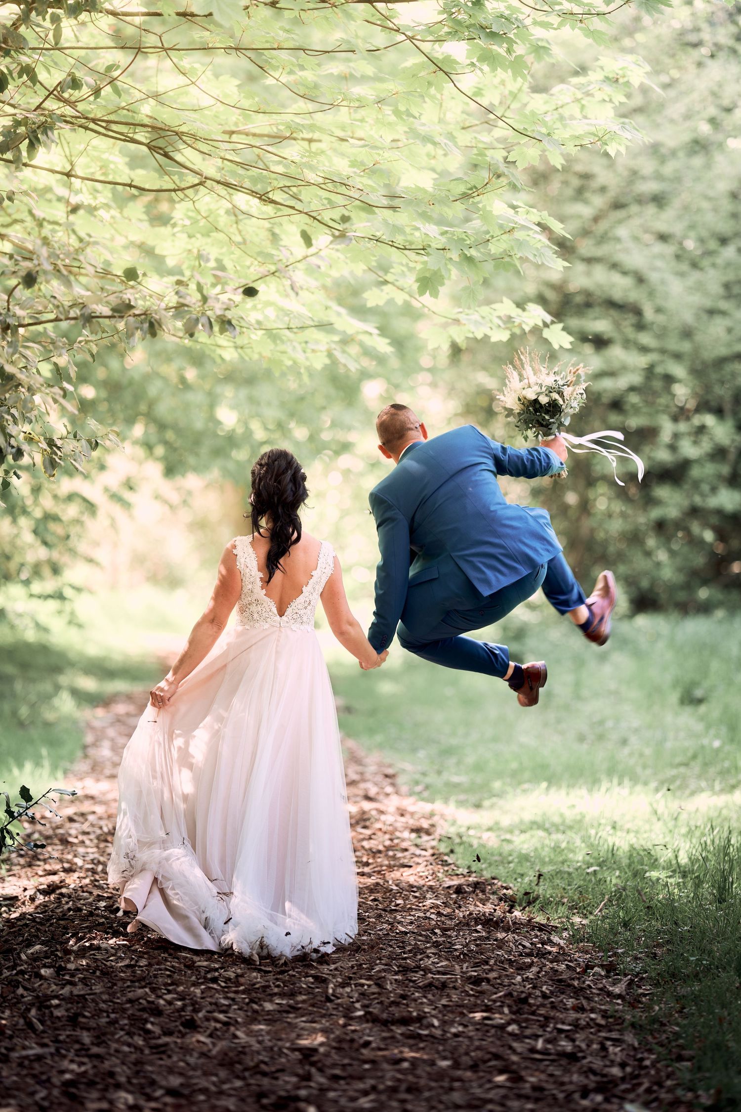 Joyful groom jumping with bouquet beside bride in white lace gown on a tree-lined path.