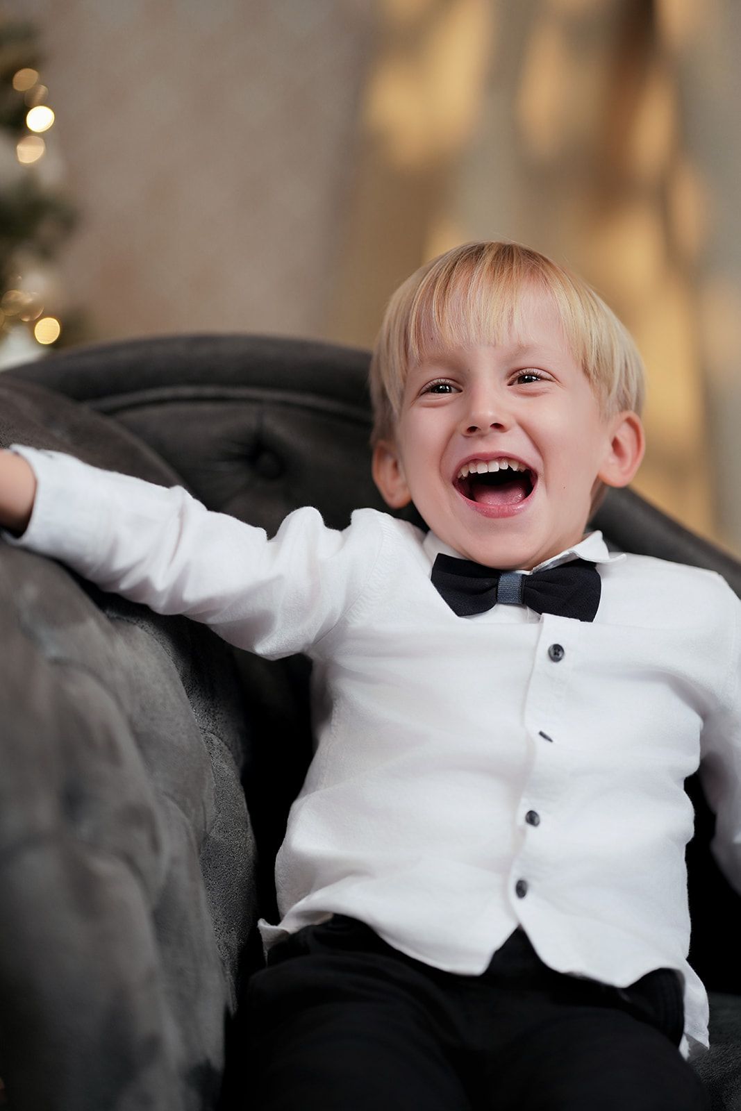 Child in formal white shirt and bowtie laughing joyfully on couch.