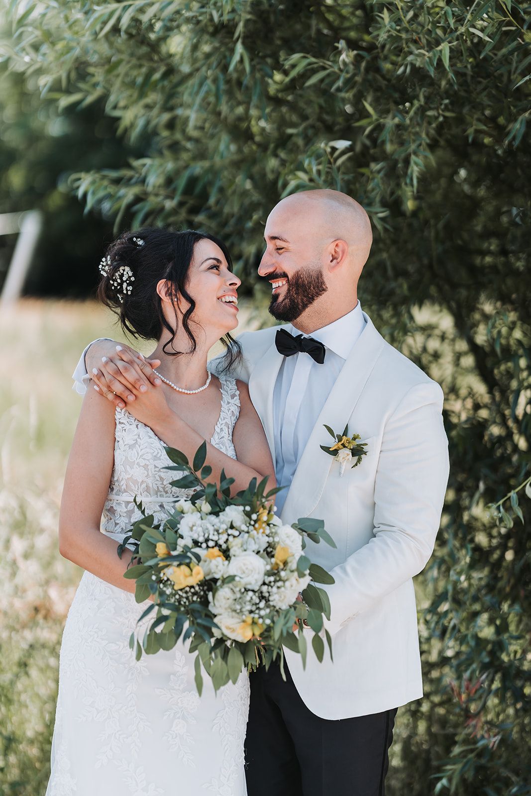 A romantic wedding portrait of a couple laughing together under trees with the bride holding a beautiful floral bouquet.