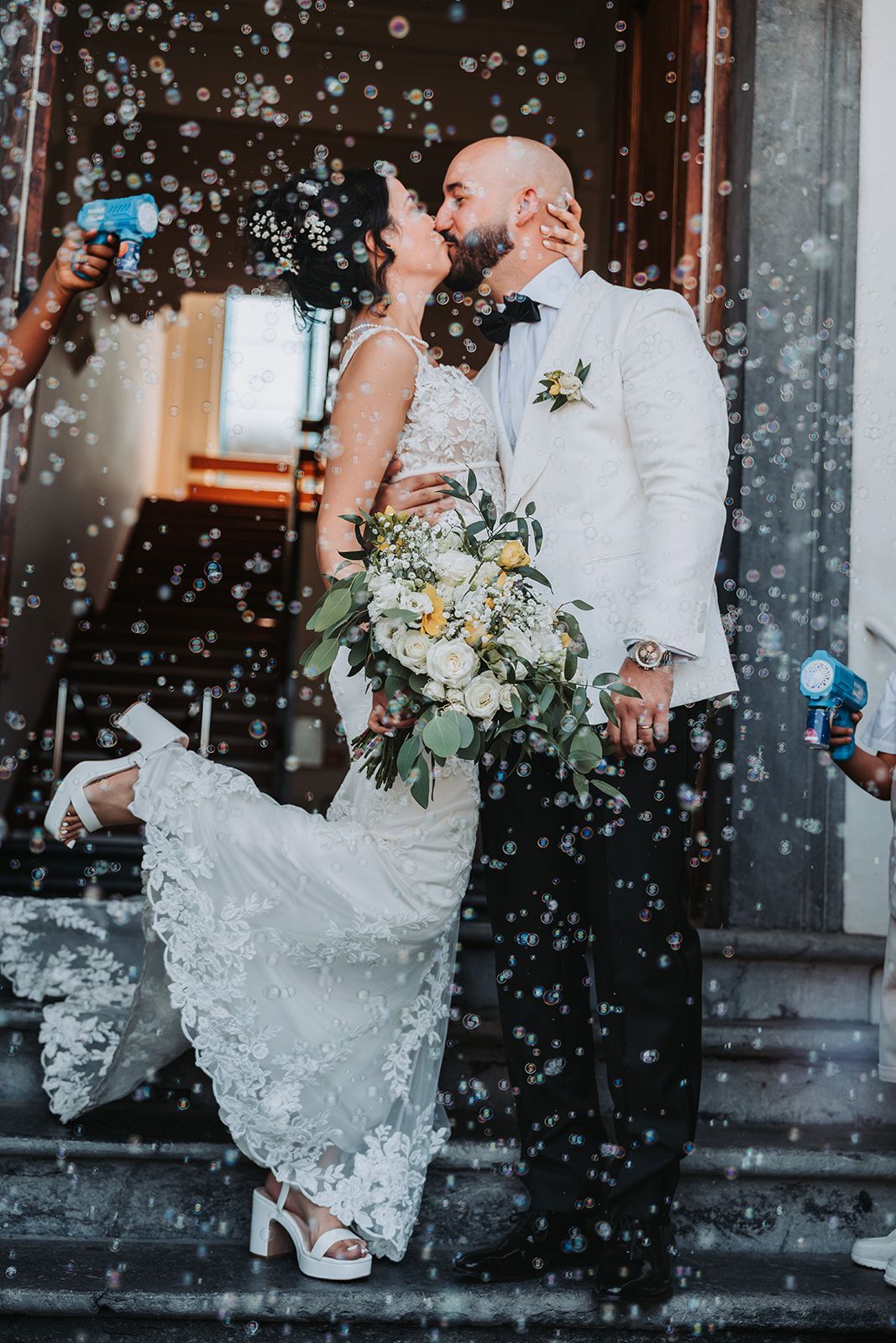 Newlyweds share a kiss while confetti falls around them on stone steps.