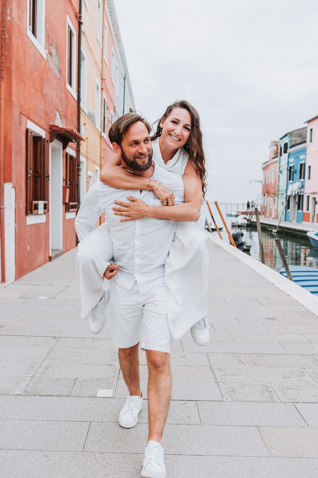 Couple in white outfits playfully running and laughing along a coral-colored building walkway.