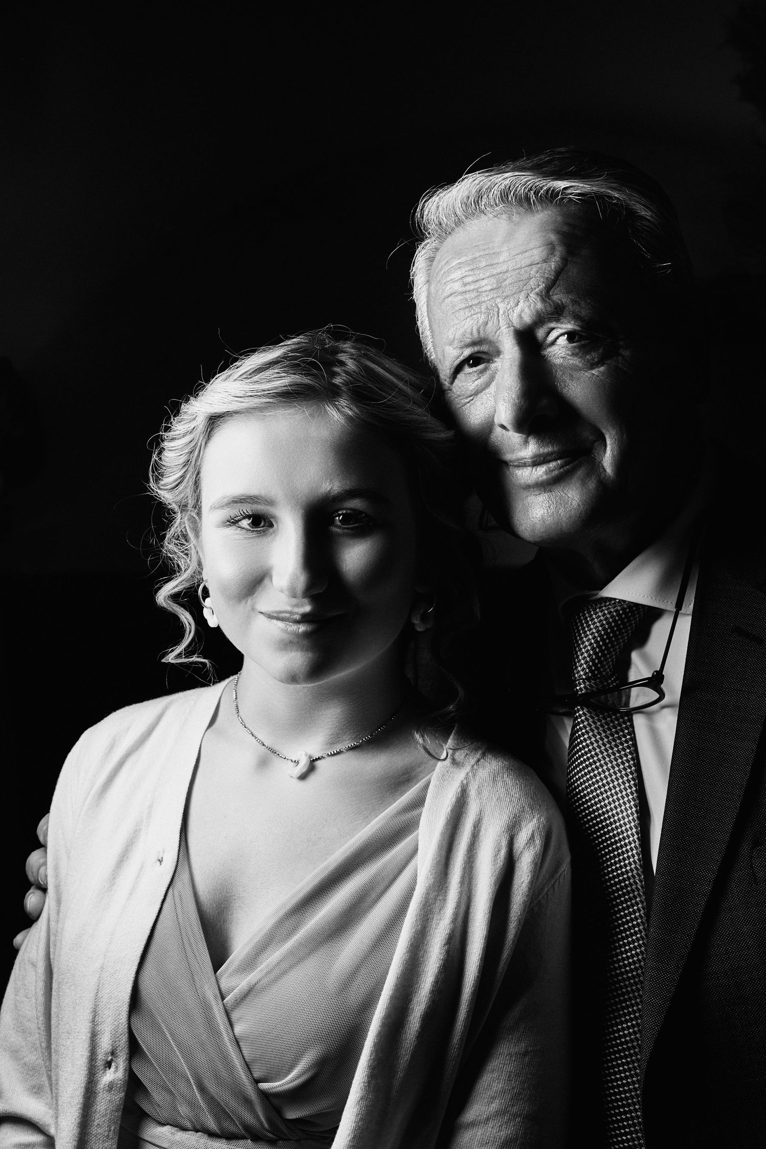 Black and white portrait of a smiling young woman and older man in a suit, posing together against a dark background.