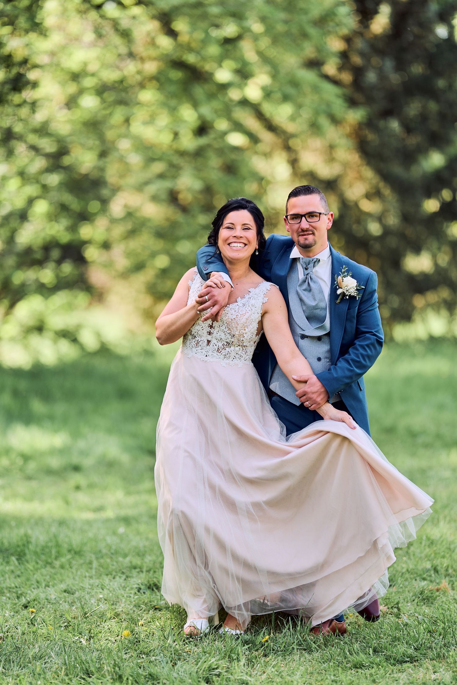 Joyful bride and groom dancing outdoors on green grass, bride in blush gown, groom in blue suit with boutonniere.