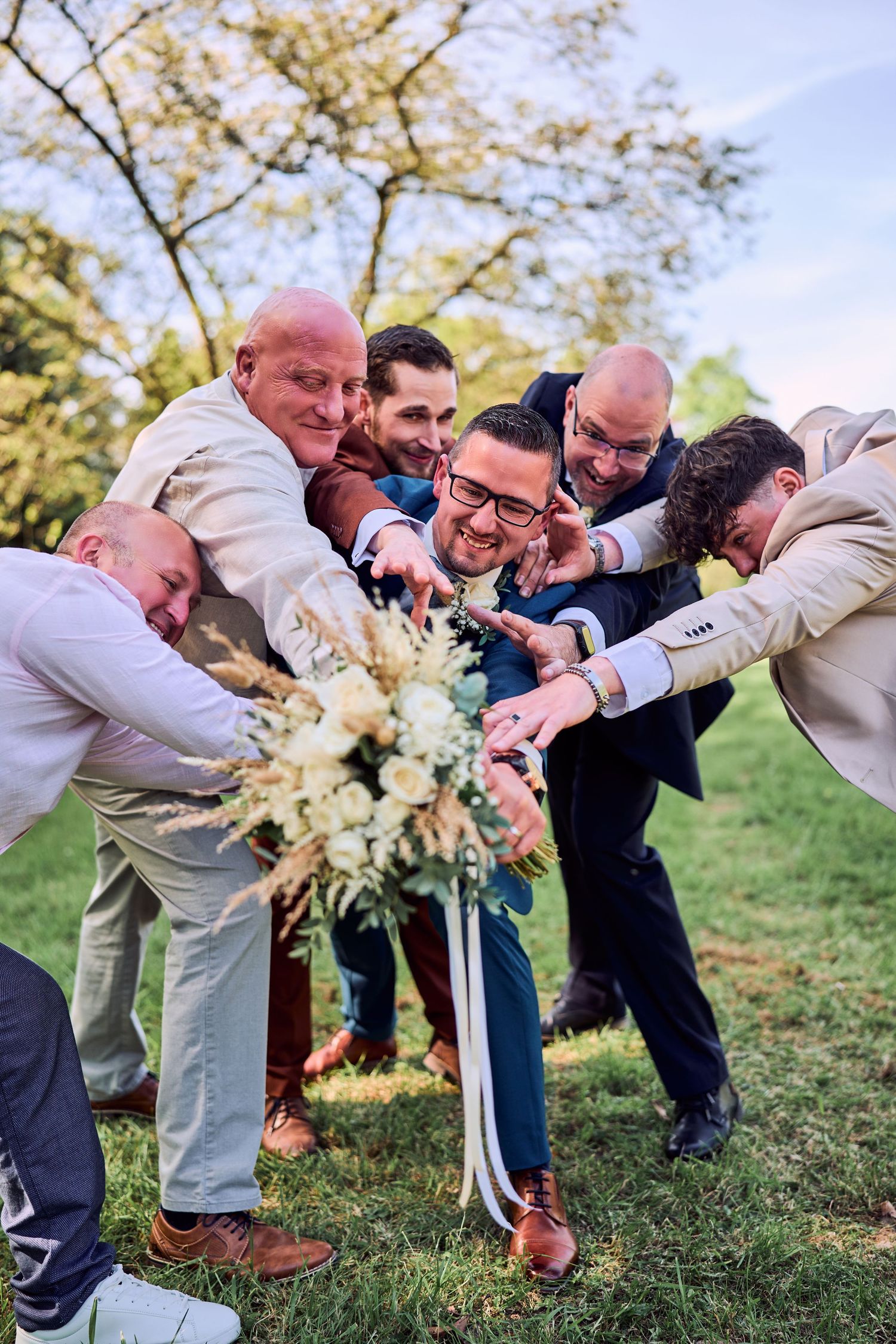 Groom and groomsmen huddle together pointing at a white floral bouquet outdoors at a wedding celebration.