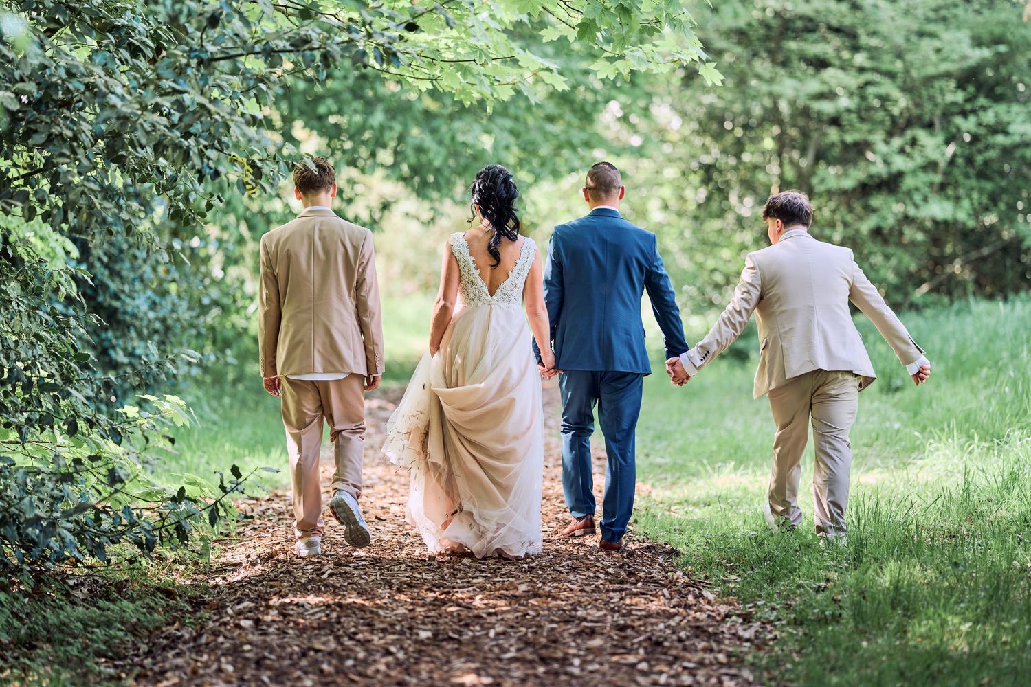 Wedding party walking along a wooded path, bride in white gown holding hands with groom in blue suit.