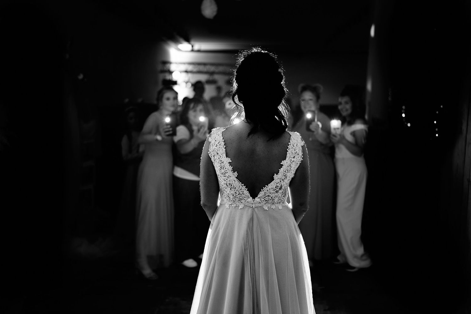 Black and white photo of bride in lace back wedding gown facing bridesmaids holding glowing phones in dark venue.