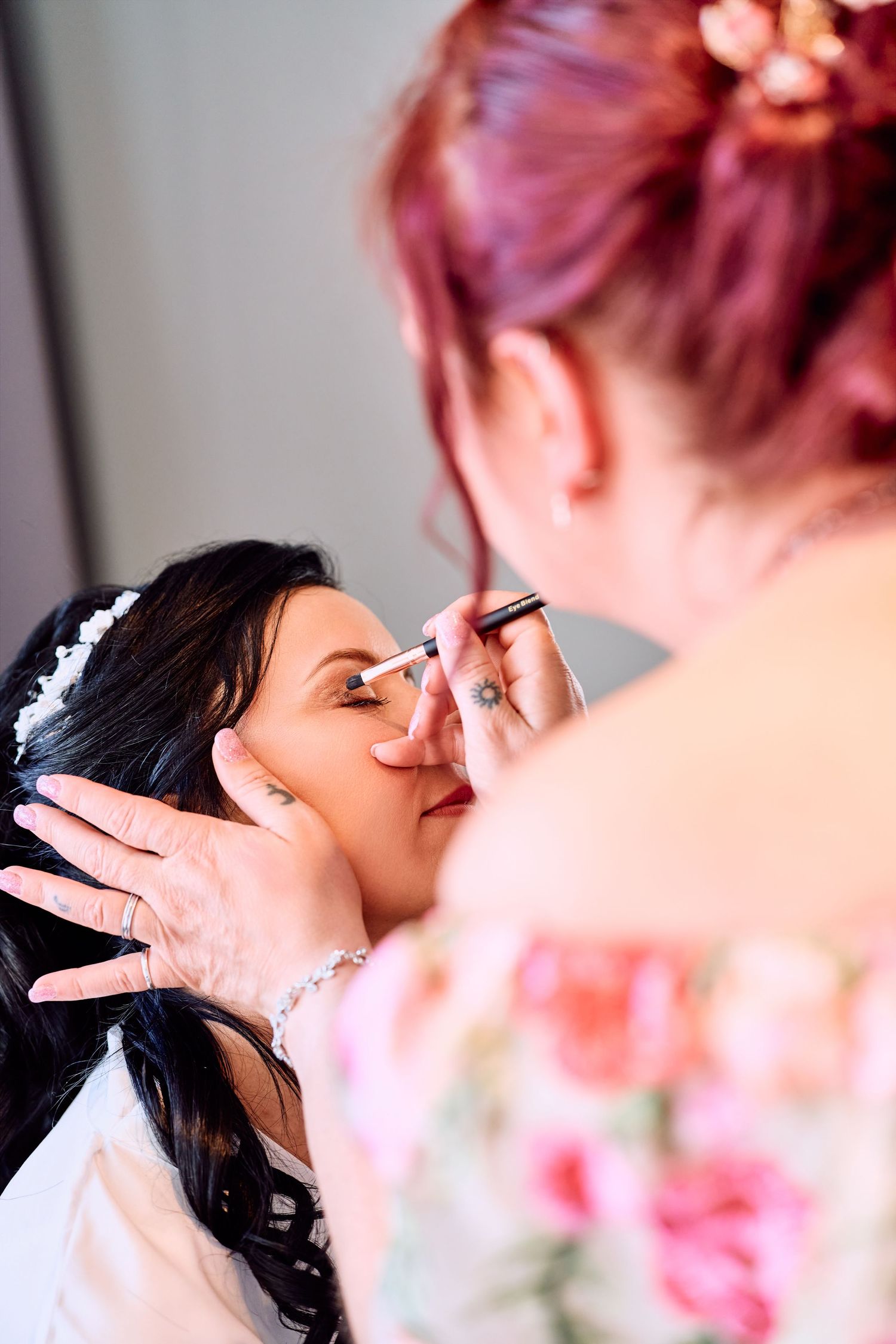 Professional makeup artist applying eye makeup to dark-haired bride wearing white robe before wedding ceremony.