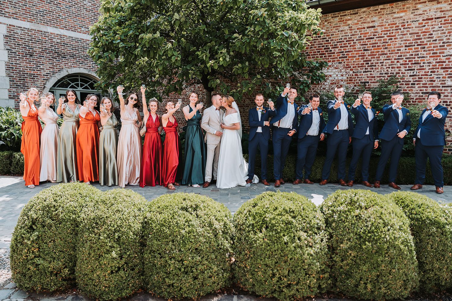Wedding party members in colorful dresses and suits pose together in an outdoor garden setting.