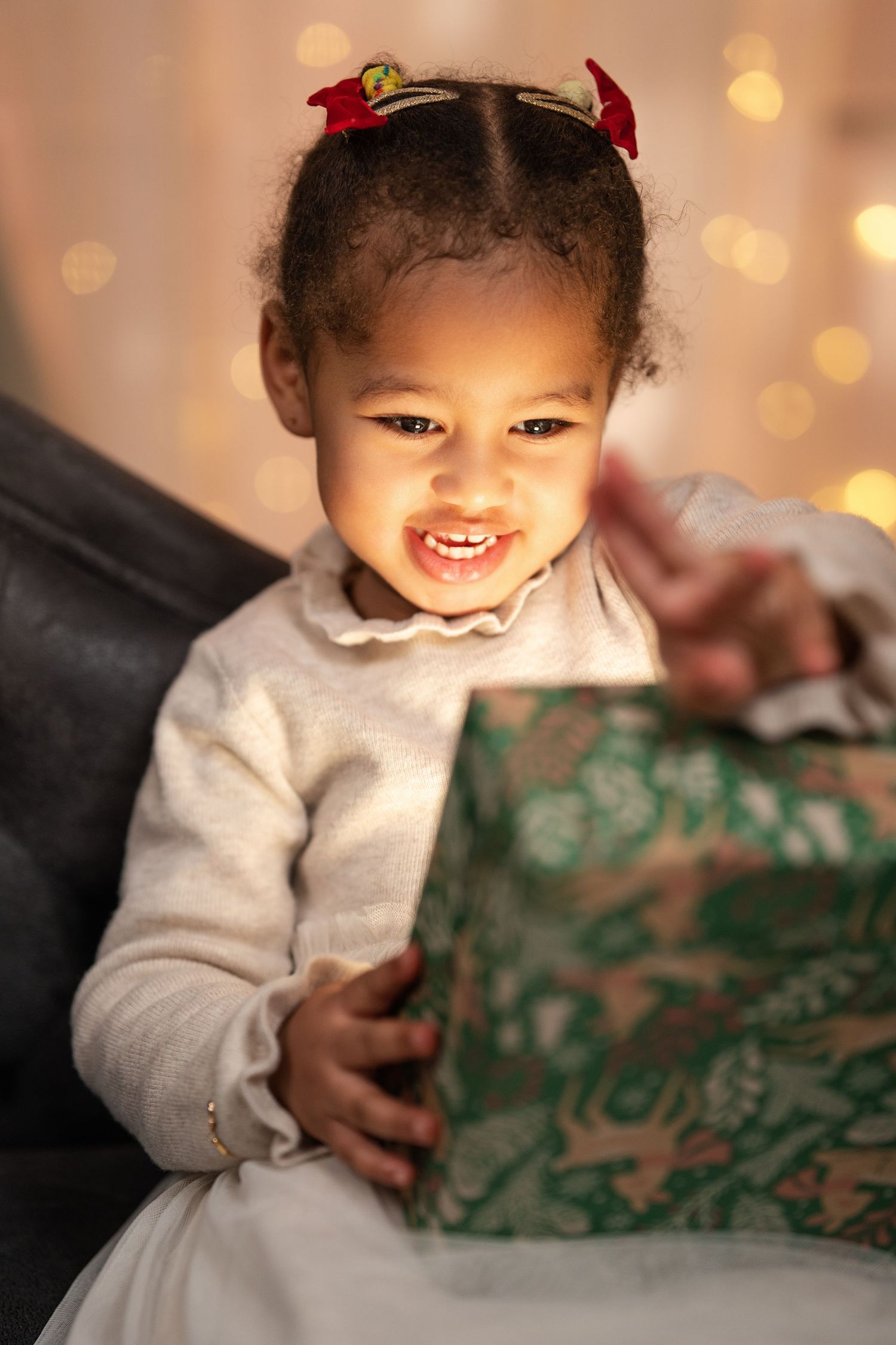 Young child in white dress holding a wrapped present with holiday lights in background.