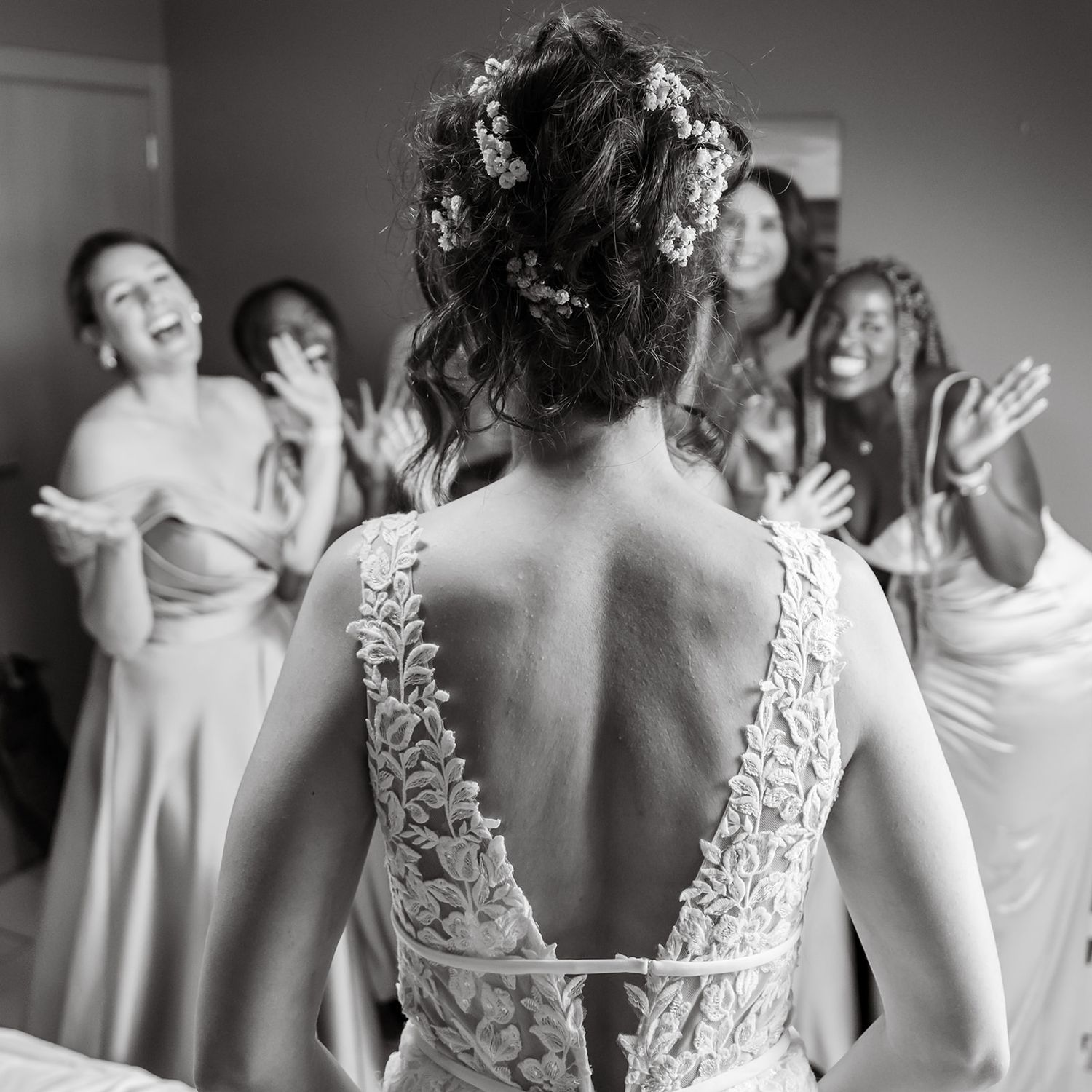 Back view of a detailed lace wedding dress while bridesmaids react with joy.
