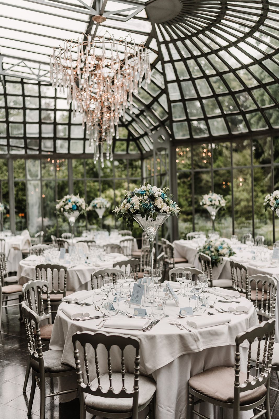 Elegant greenhouse wedding reception setup with crystal chandelier and floral centerpieces.