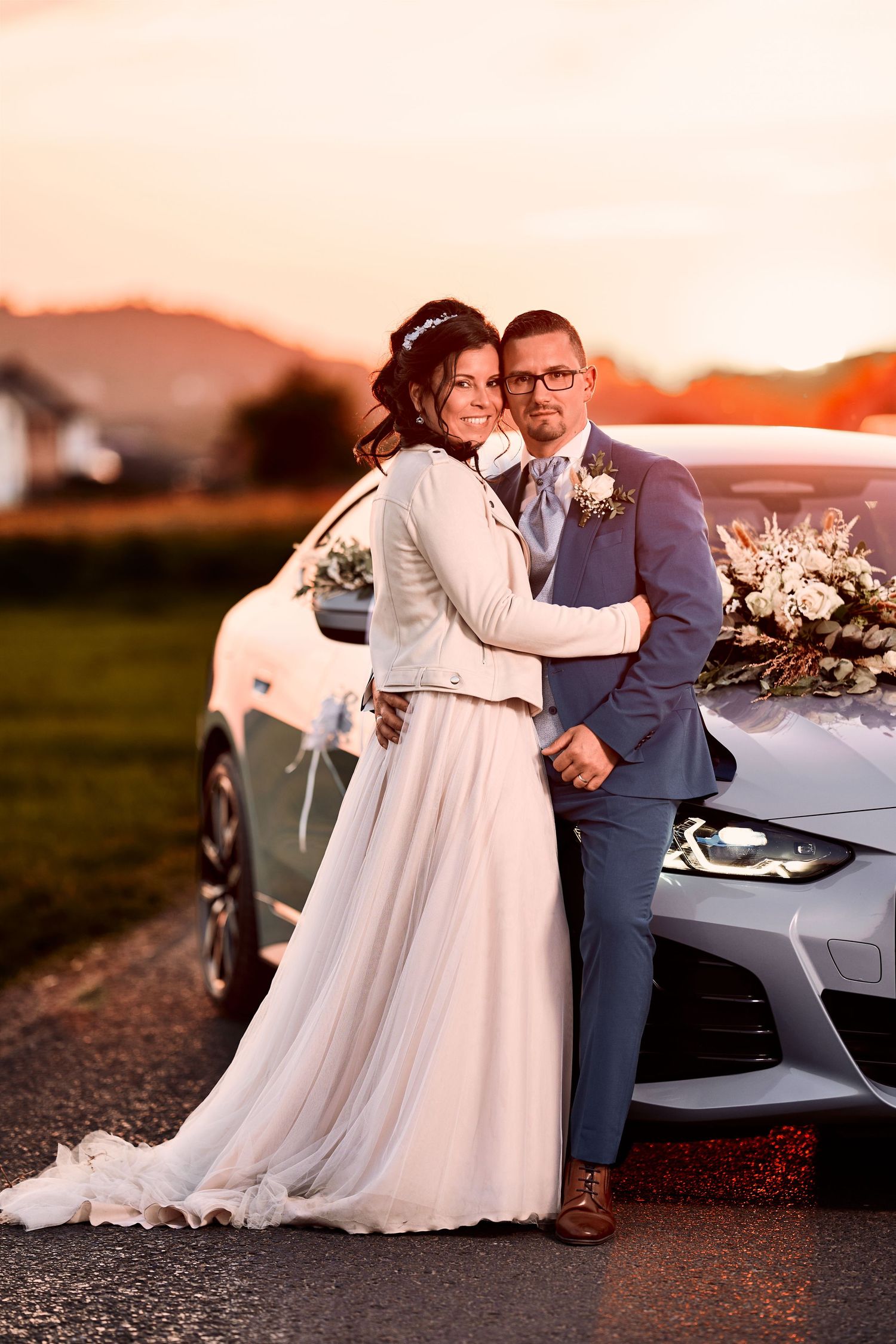Bride and groom pose together leaning against a decorated car during a golden sunset wedding photo.