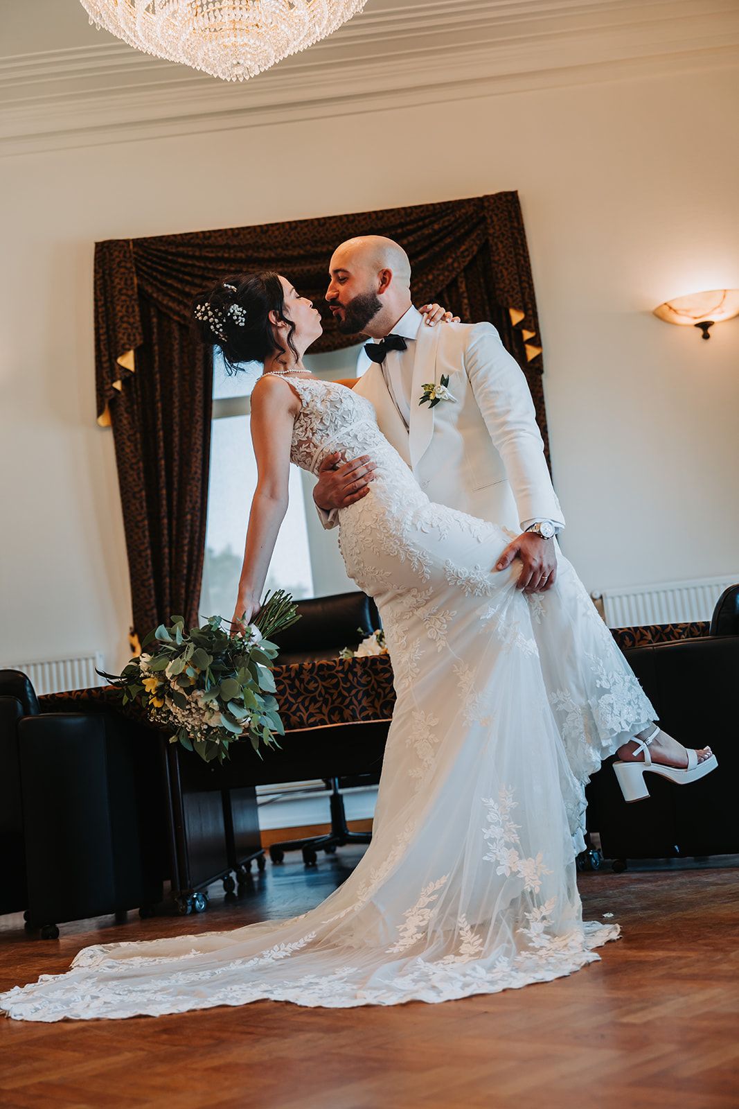 Romantic wedding portrait in front of a dark curtain with an elegant chandelier overhead.
