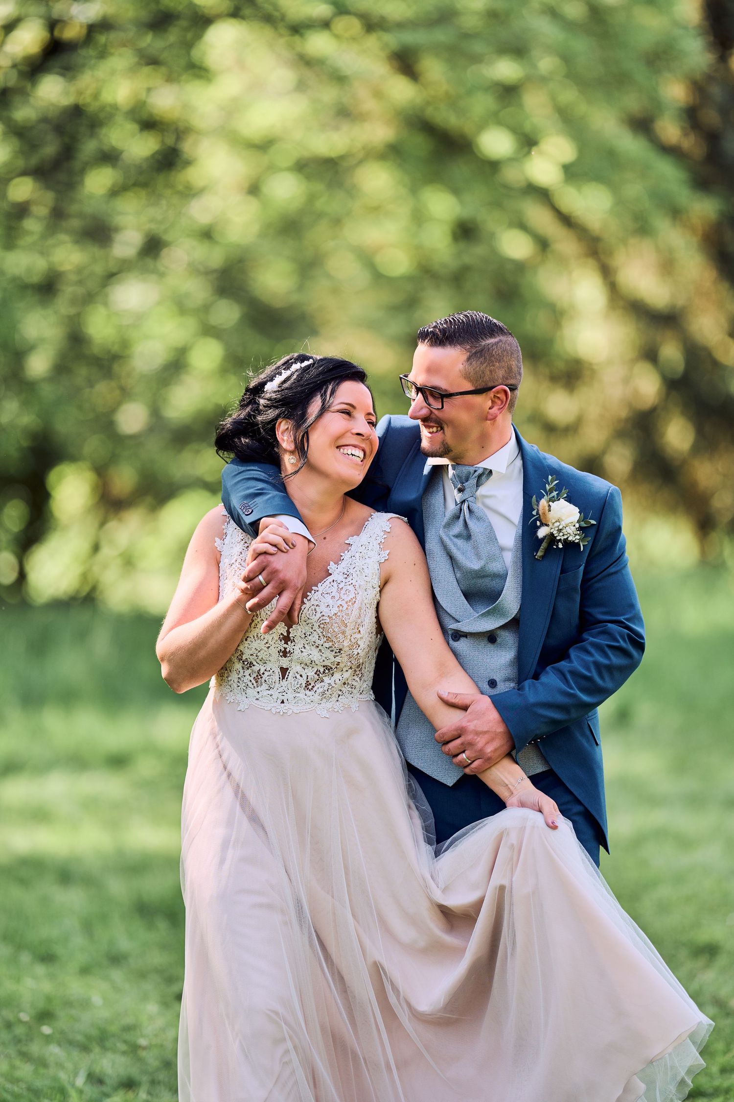 Happy bride and groom embracing outdoors in lush green park, bride in beige lace gown, groom in blue suit.