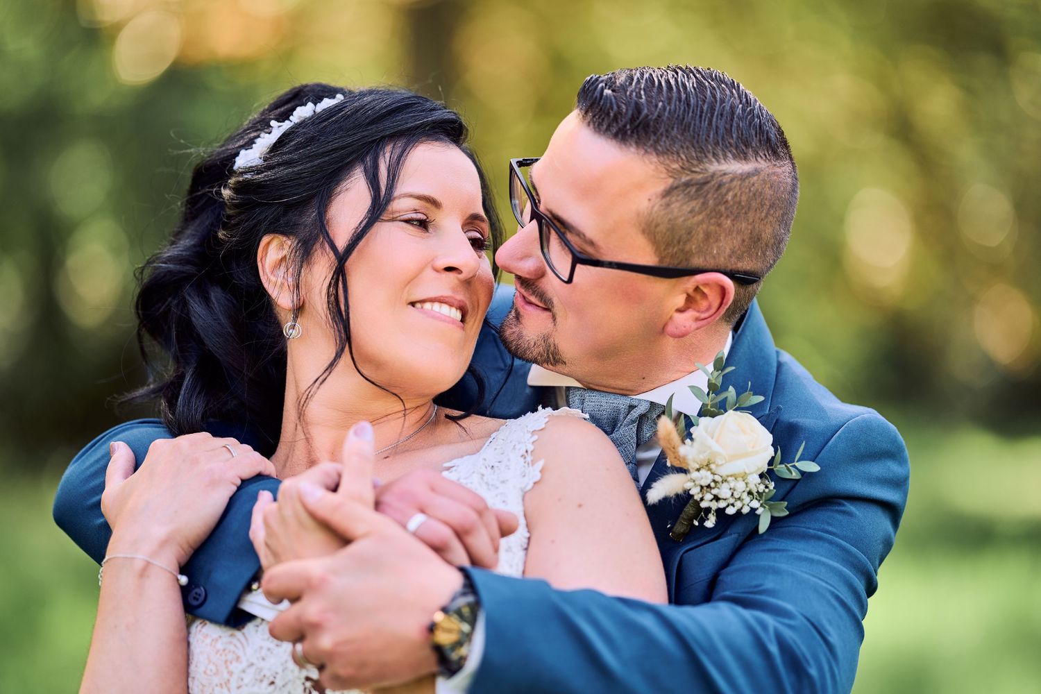 Happy newlyweds embrace outdoors, bride in white lace gown and groom in blue suit with glasses, sharing a joyful moment.