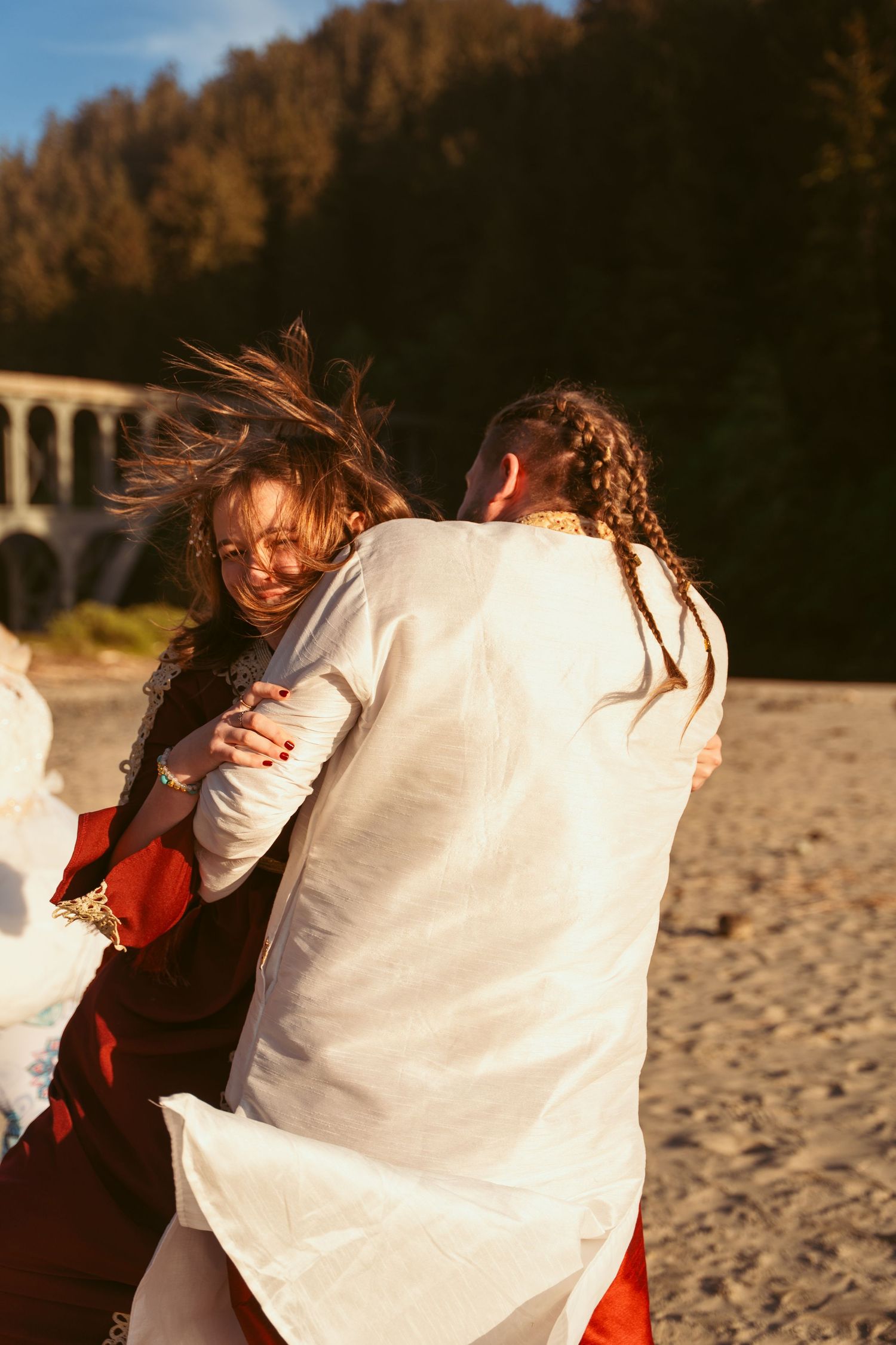 Two people in flowing red and white attire embrace while dancing on a scenic beach at golden hour.