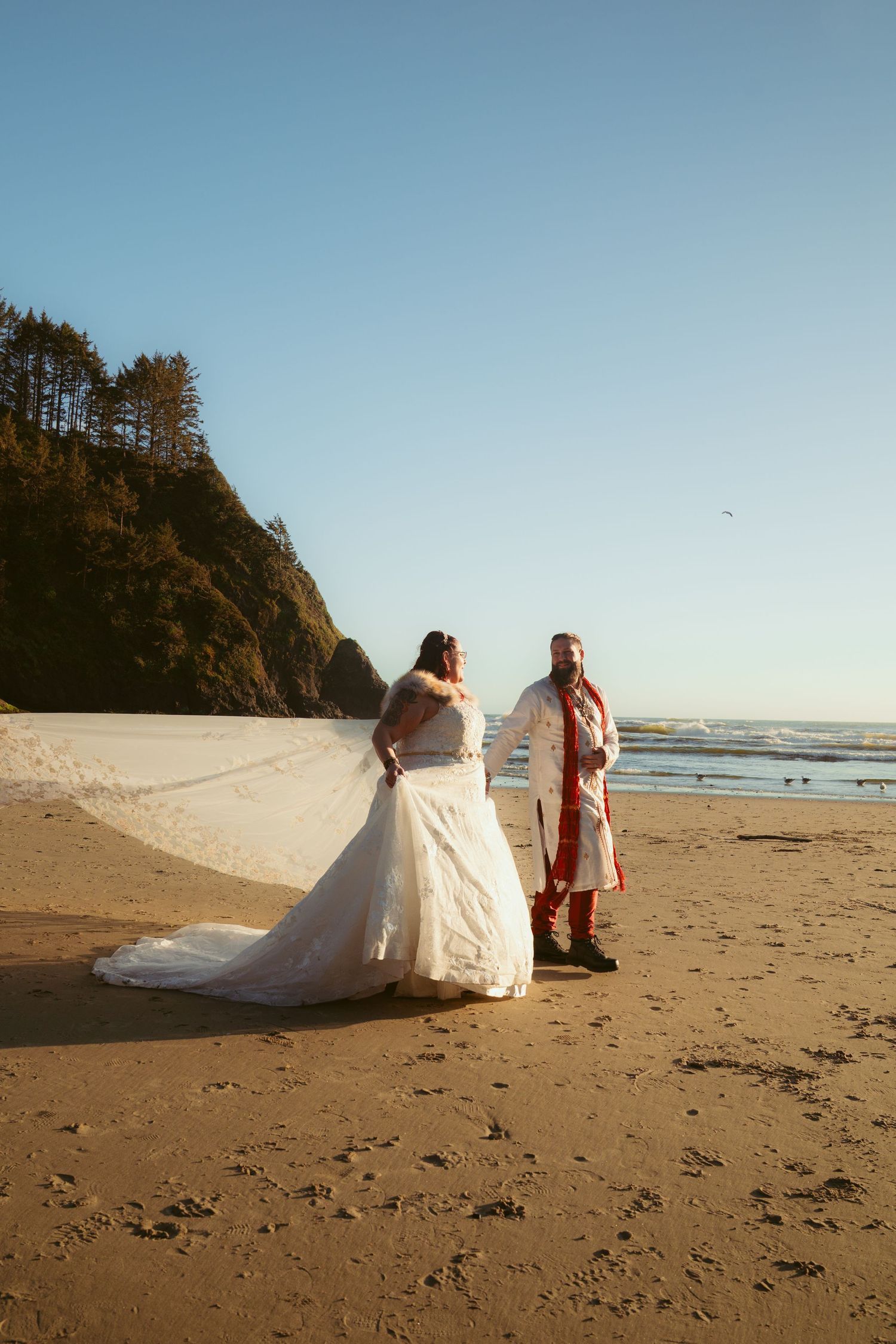 A couple in wedding attire walks along a scenic beach with cliffs in the background.