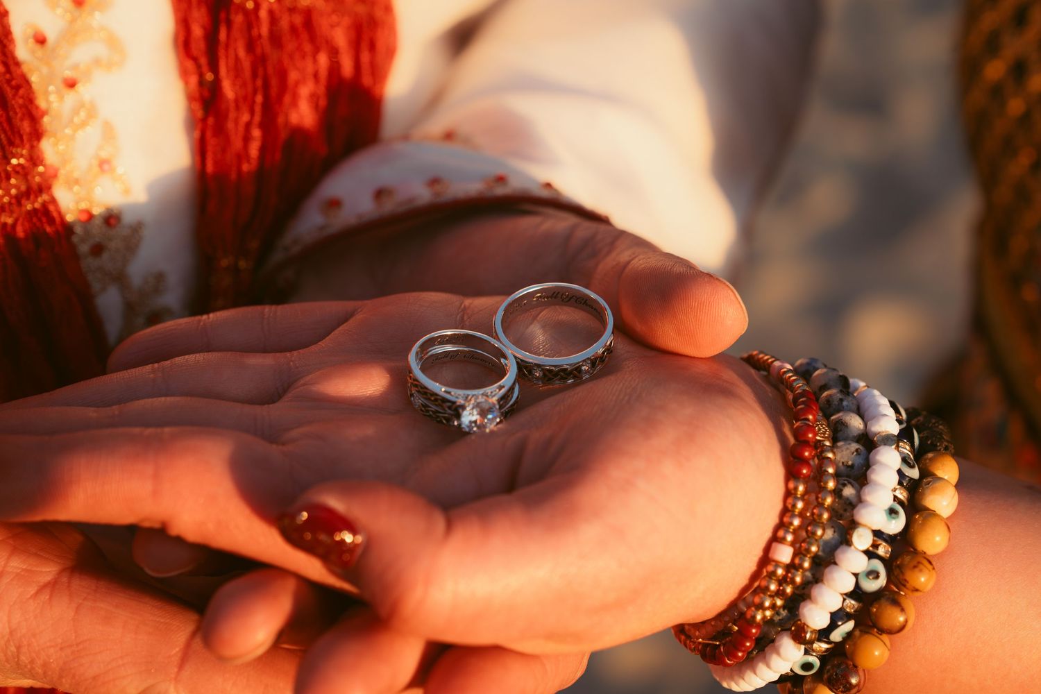 Wedding rings displayed on hands with warm lighting and beaded bracelets.