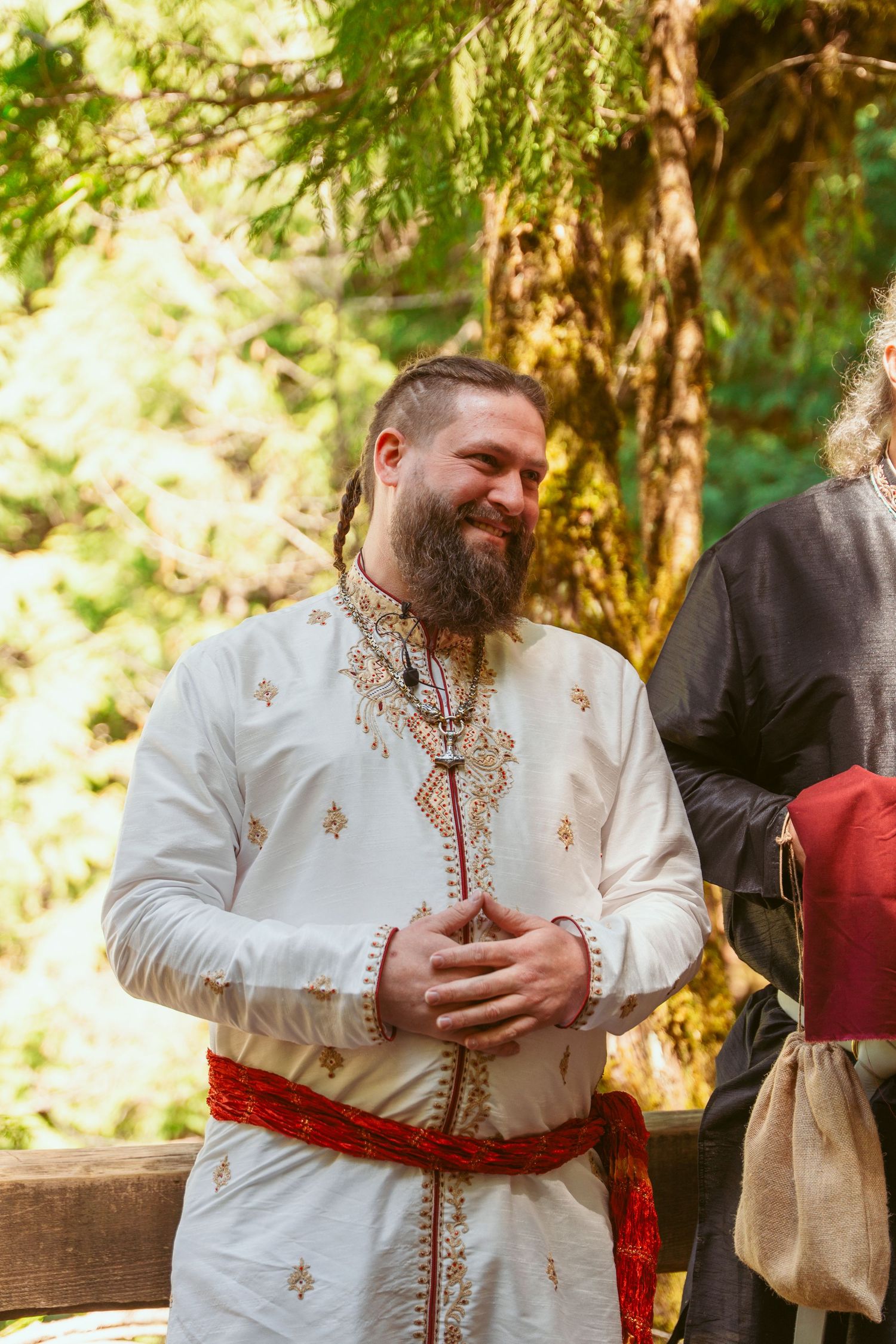 Person in traditional white medieval clothing with embroidery and red sash during forest ceremony.