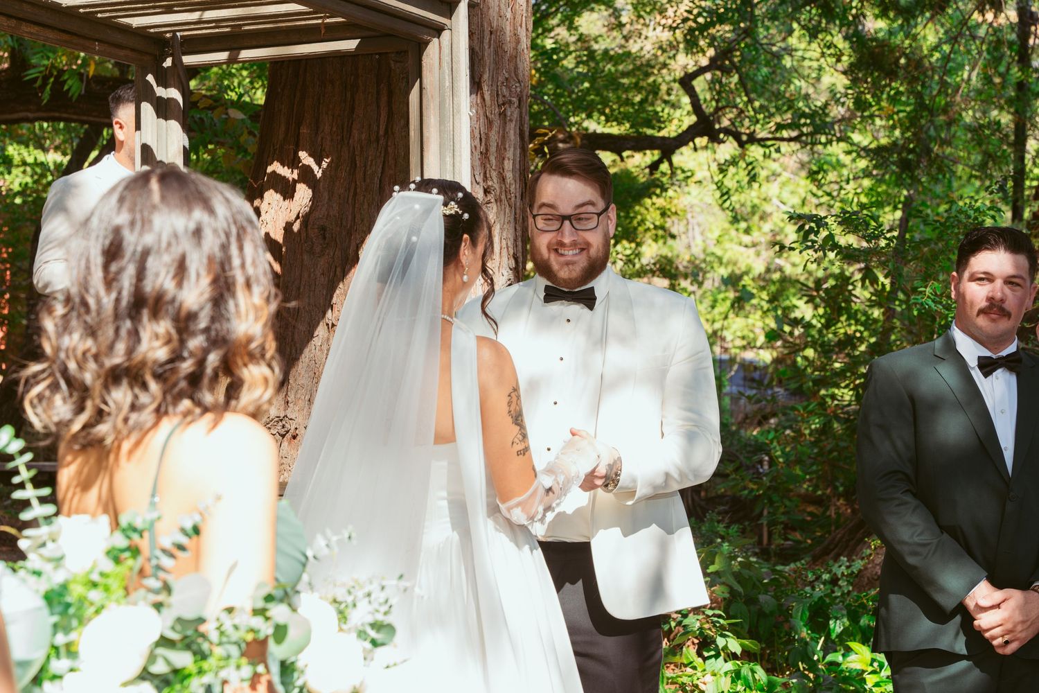 Wedding ceremony taking place under rustic wooden gazebo with natural greenery.