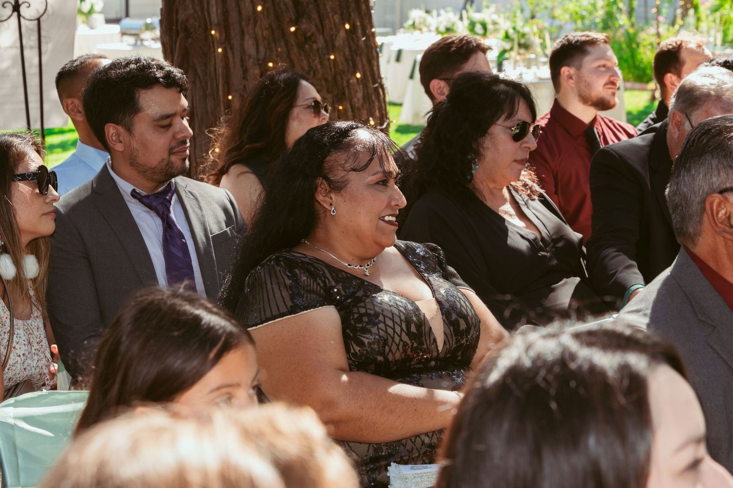 Wedding guests seated outdoors watch ceremony under string lights and tall trees.