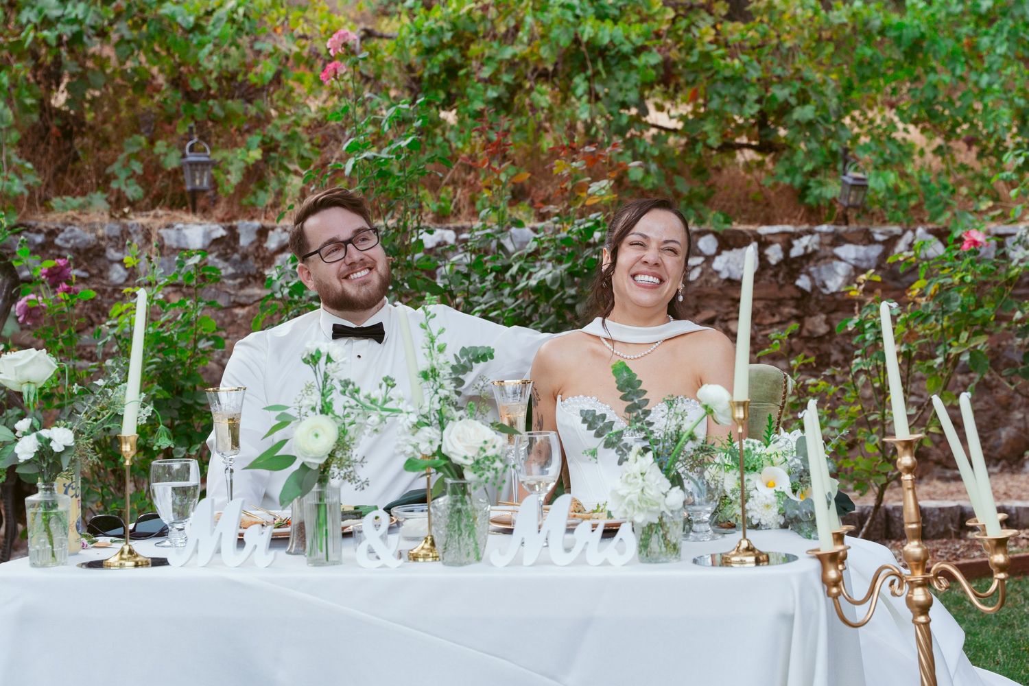 Newlyweds seated at elegant white reception table with greenery and candelabra decorations.