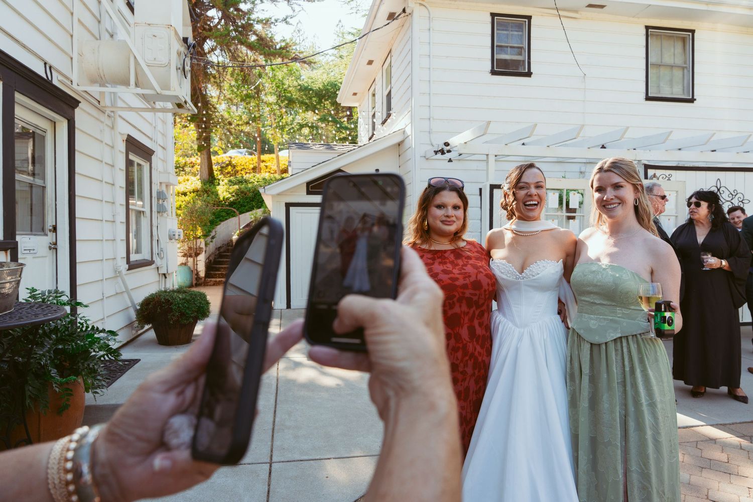 Wedding party members pose and dance outside a white building during daytime celebrations.