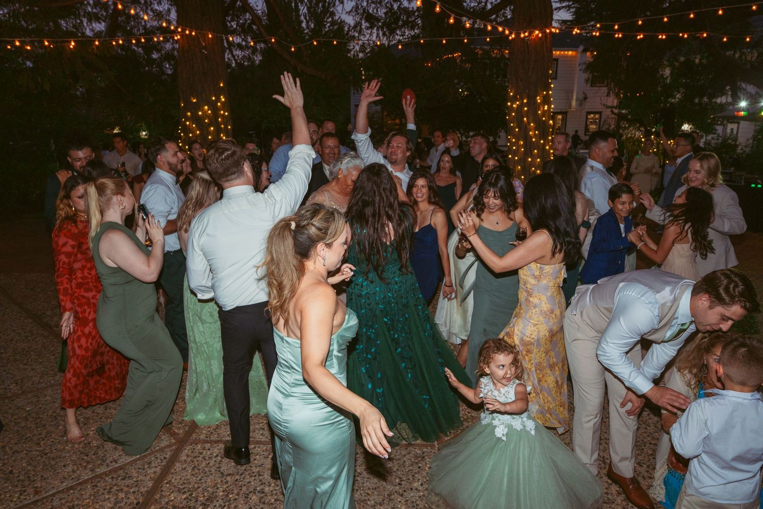 Wedding celebration on an outdoor dance floor illuminated by warm string lights as guests dance and socialize.