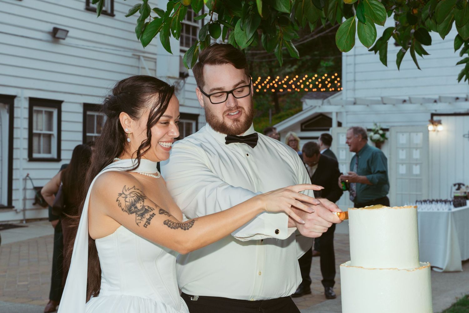 A joyful cake cutting ceremony at an outdoor wedding with a white house in the background.
