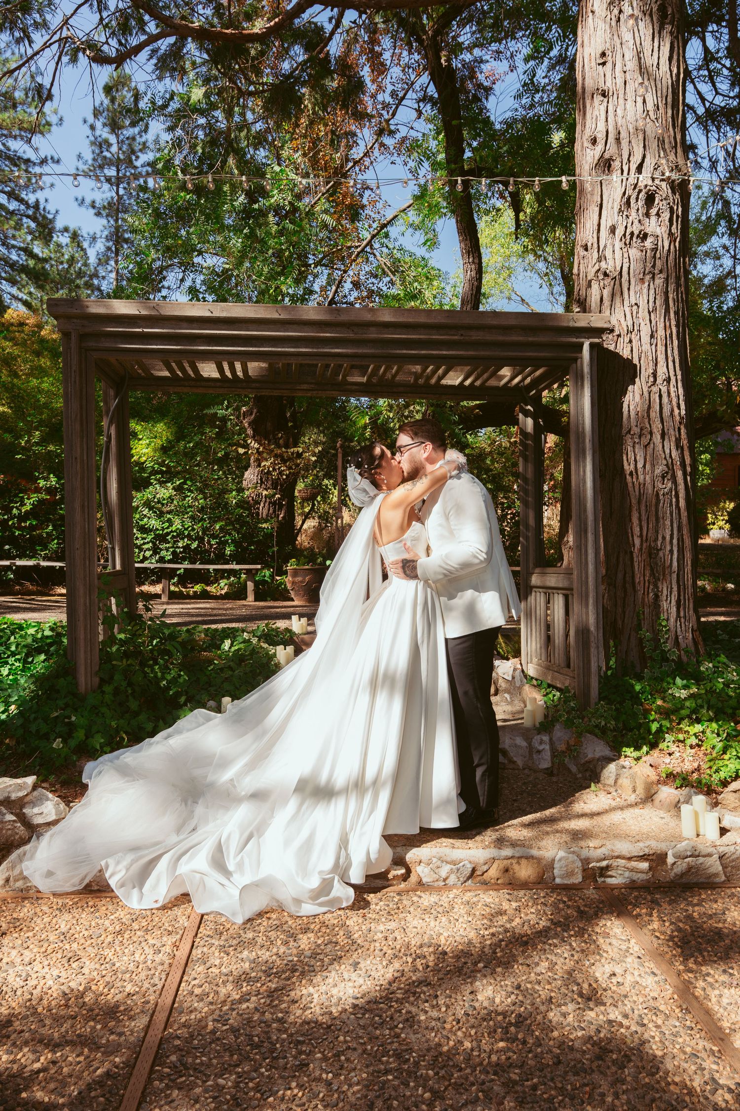 A romantic wedding portrait of a couple embracing under a wooden pergola surrounded by tall trees.