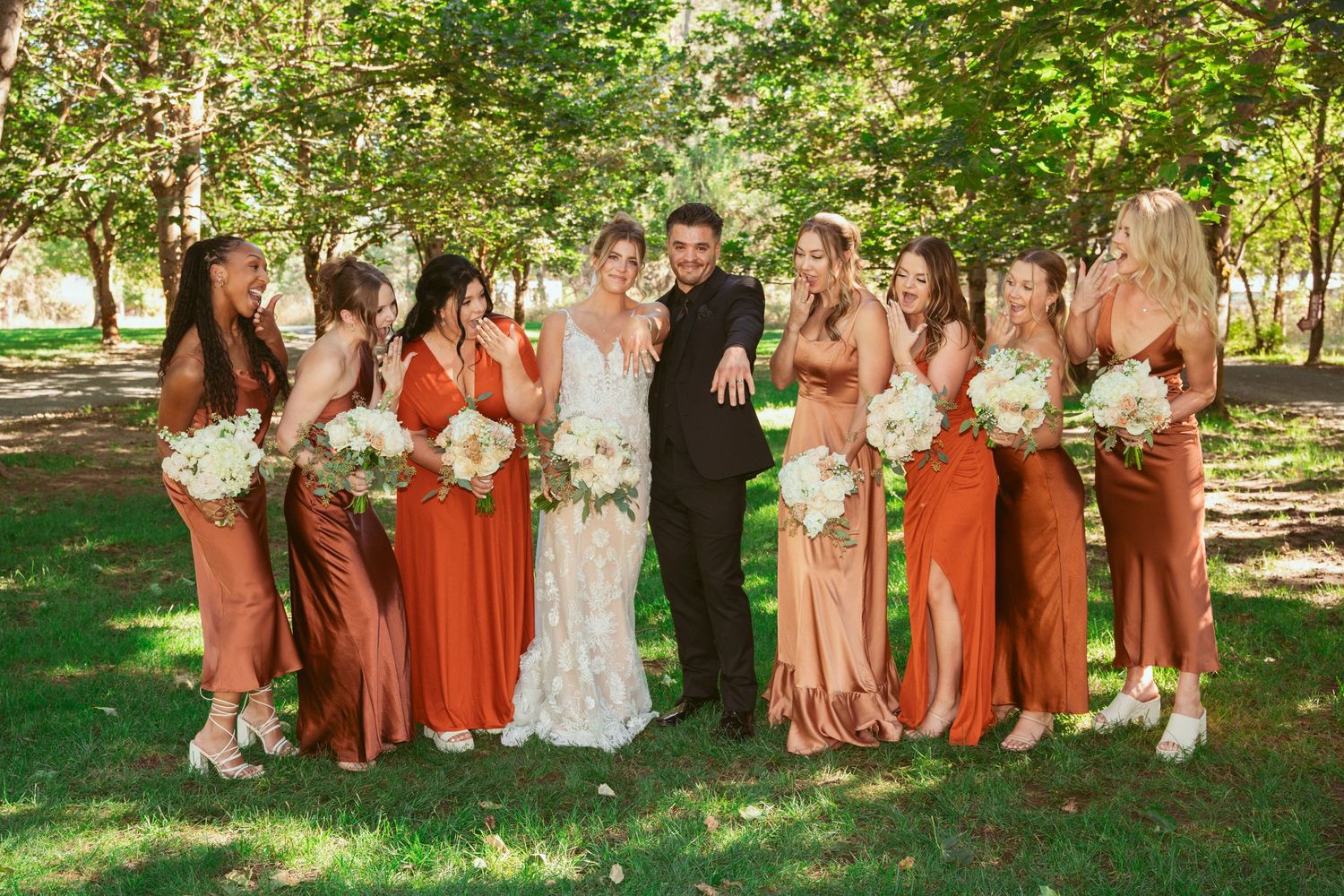 Wedding party in rust-colored bridesmaid dresses poses together on grass under trees.