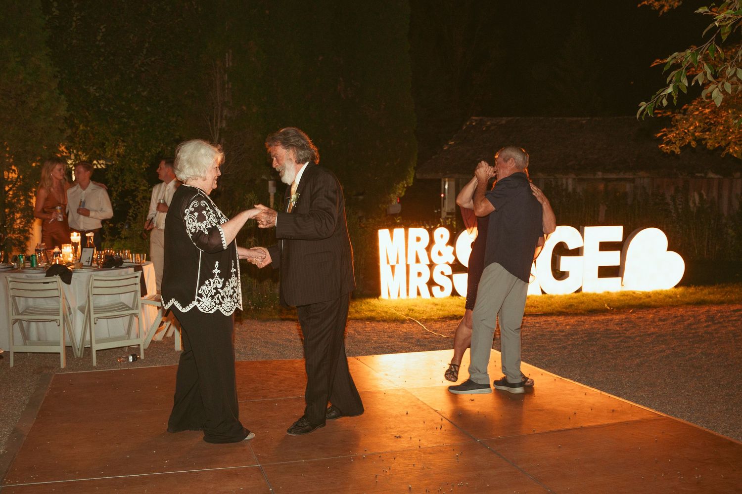 Wedding party members dancing near illuminated signage at nighttime outdoor celebration.