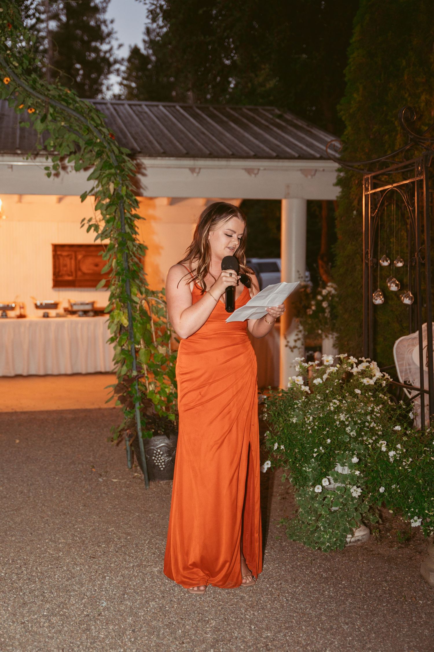 Woman in orange formal dress reads from paper at outdoor evening event with decorated venue.