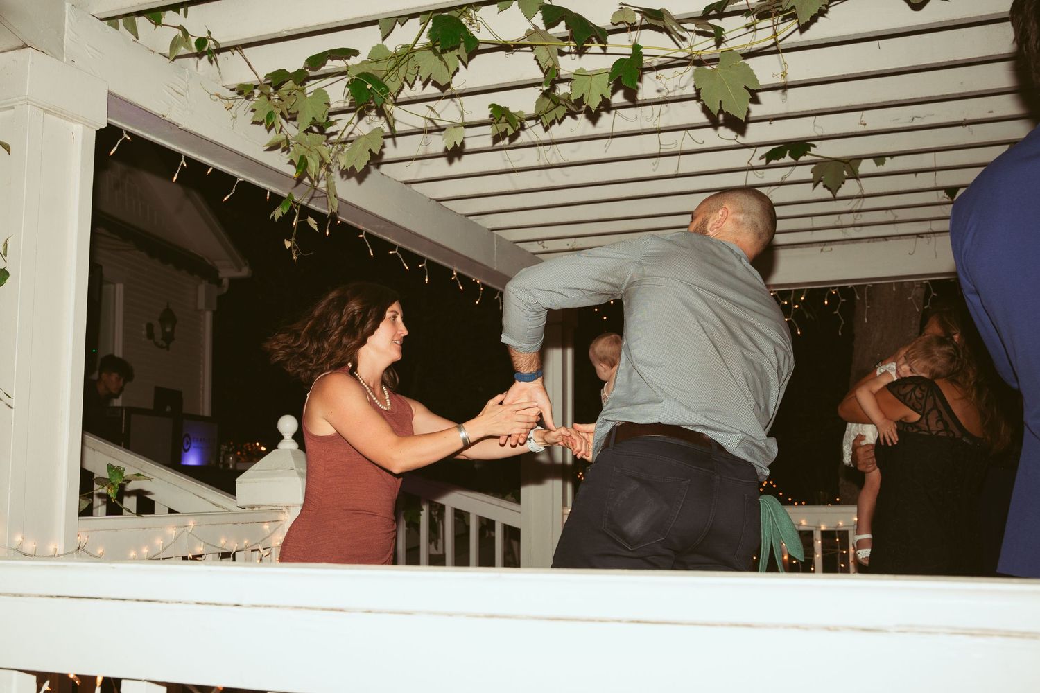 Wedding guests mingle and dance under a white pergola at an outdoor evening reception.