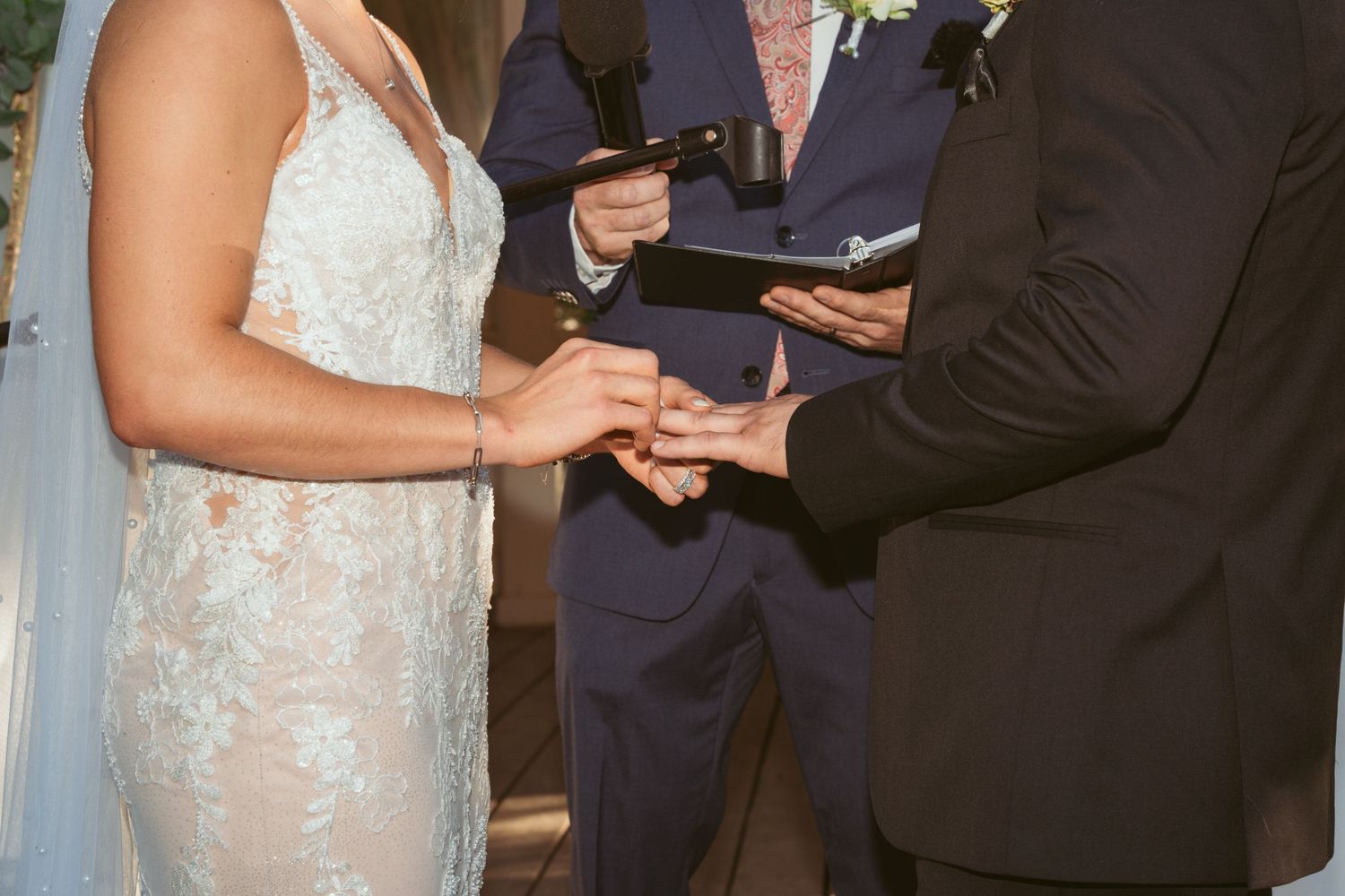 A couple exchanges rings during their wedding ceremony while an officiant in a dark suit presides over the moment.