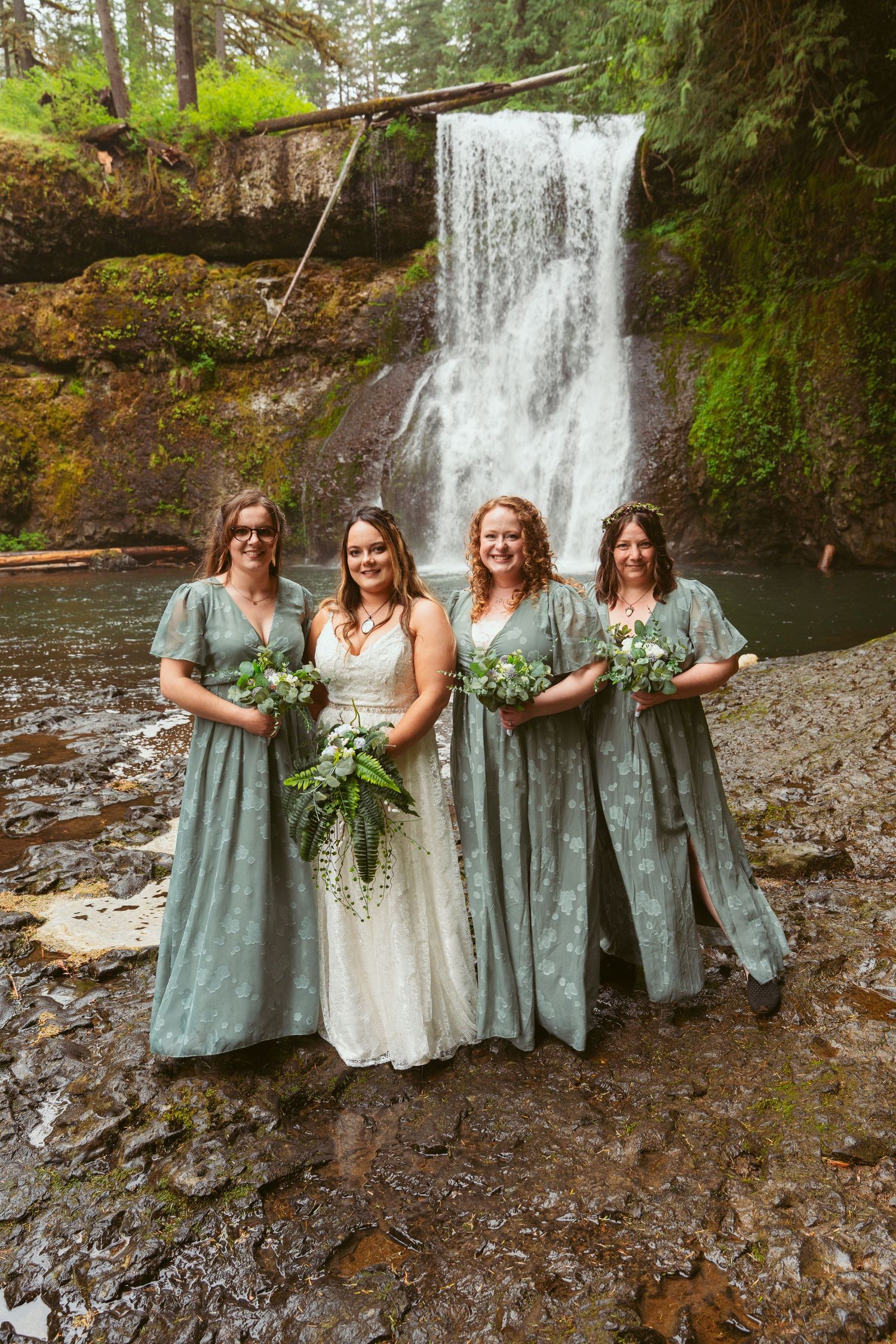 Wedding party in sage and white attire gathered for photos at a scenic forest waterfall location.