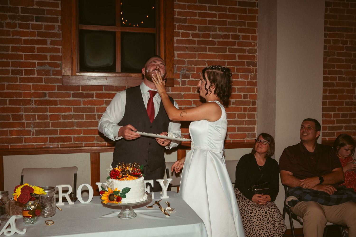 Wedding couple sharing cake at their reception with guests watching in the background.