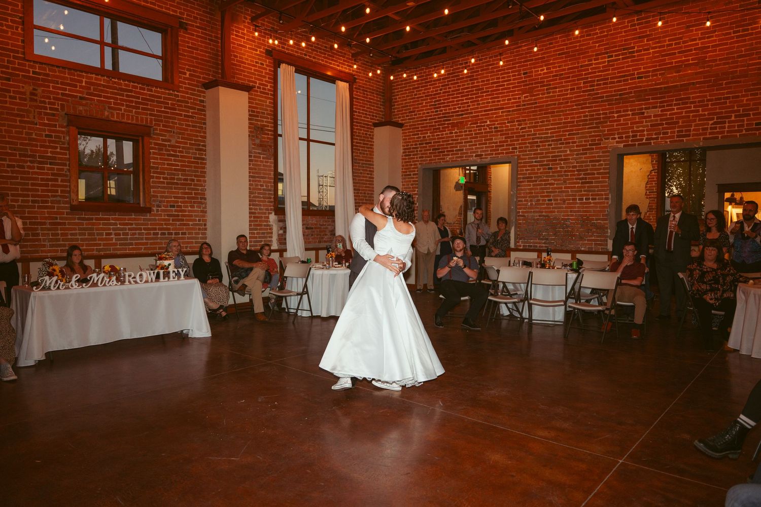 Wedding couple dancing in an industrial-style venue with exposed brick walls and string lights overhead.