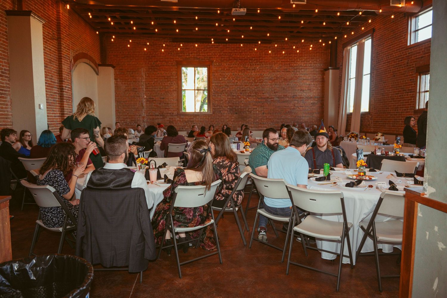 Wide view of wedding reception hall with exposed brick walls and guests seated at round tables.