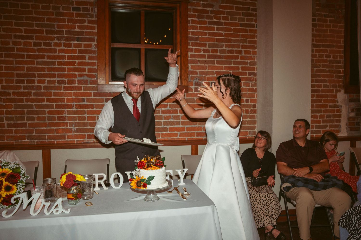 Playful cake-feeding moment between newlyweds at their reception with brick wall backdrop.