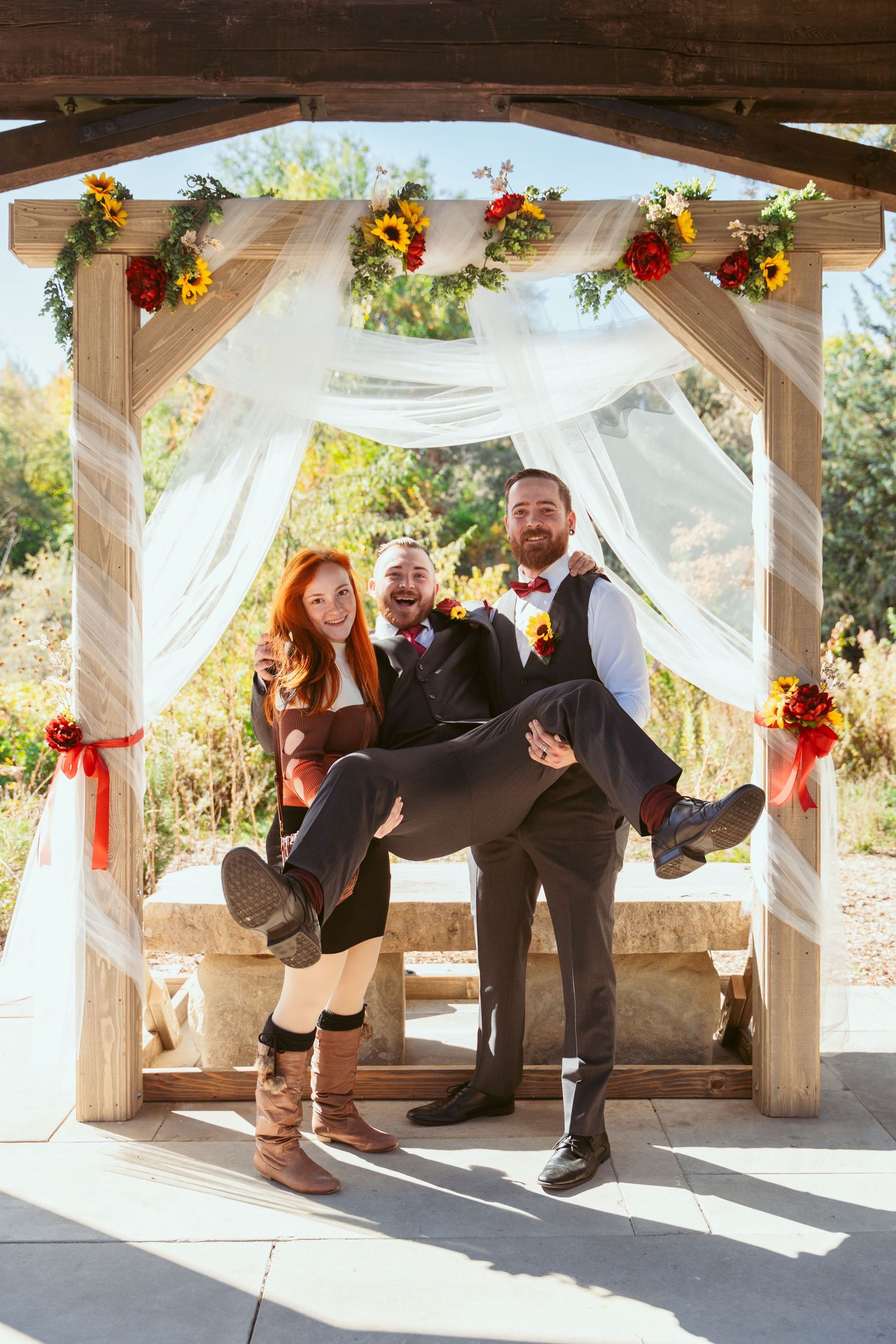 Wedding party members kick their legs up in synchronized pose under decorated outdoor pavilion.